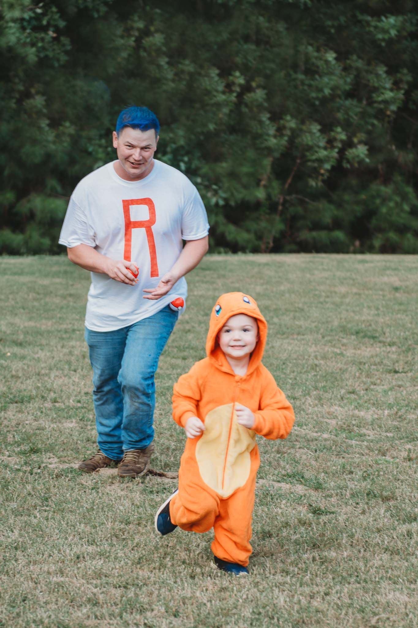 A person wearing a gray shirt with blue hair follows a small child in a bright orange hooded jumpsuit on a grassy field.
