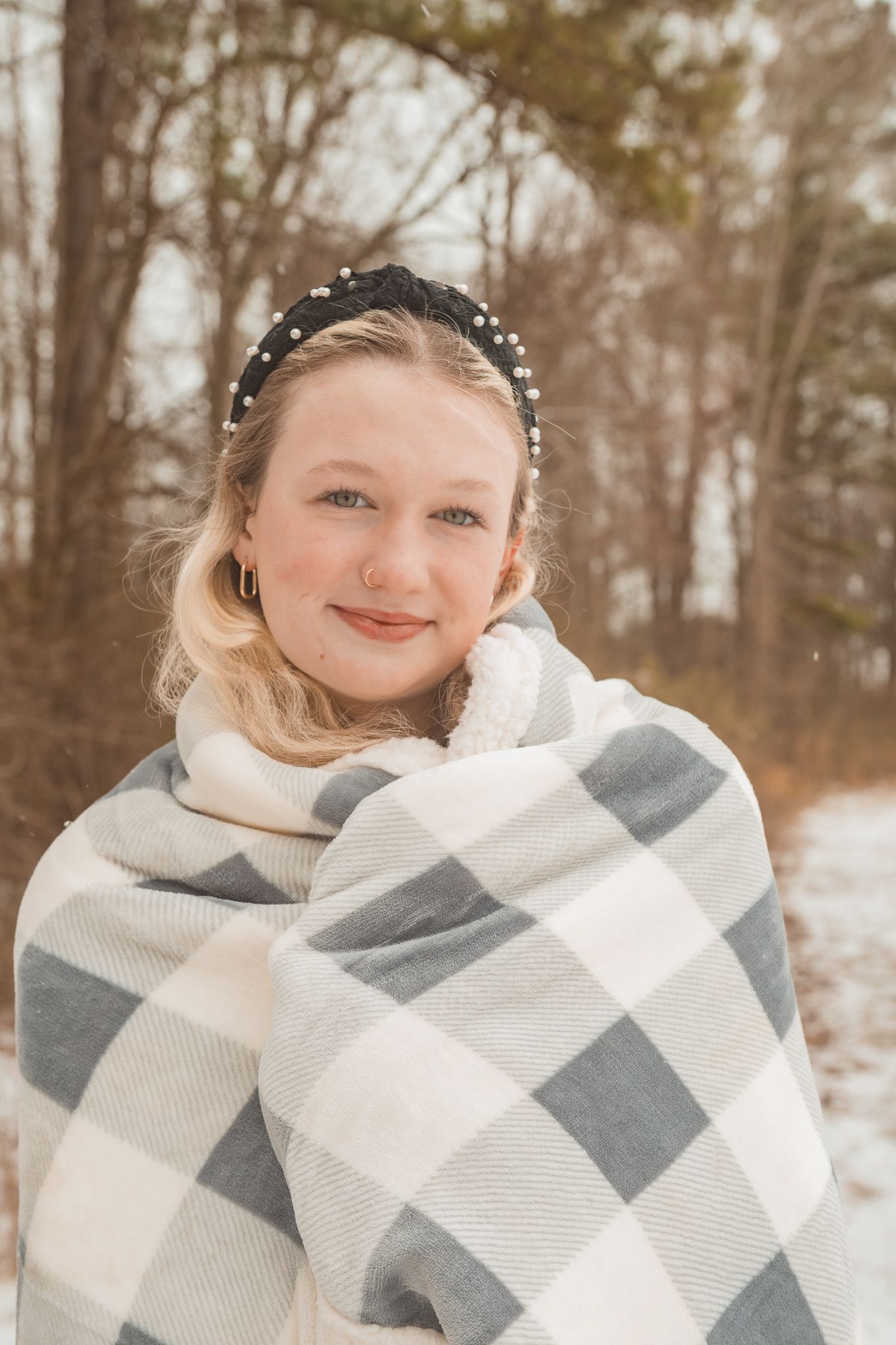 Someone wrapped in a cozy gray and white plaid blanket smiles outdoors in a snowy winter setting.