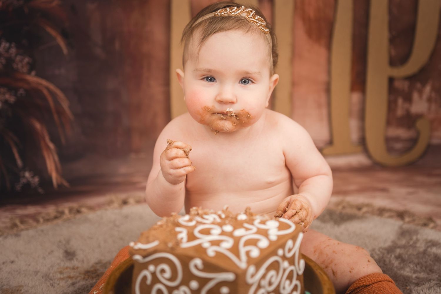 Small baby sits on rug with white swirled designs wearing a brown floral patterned bottom in warm-toned setting.