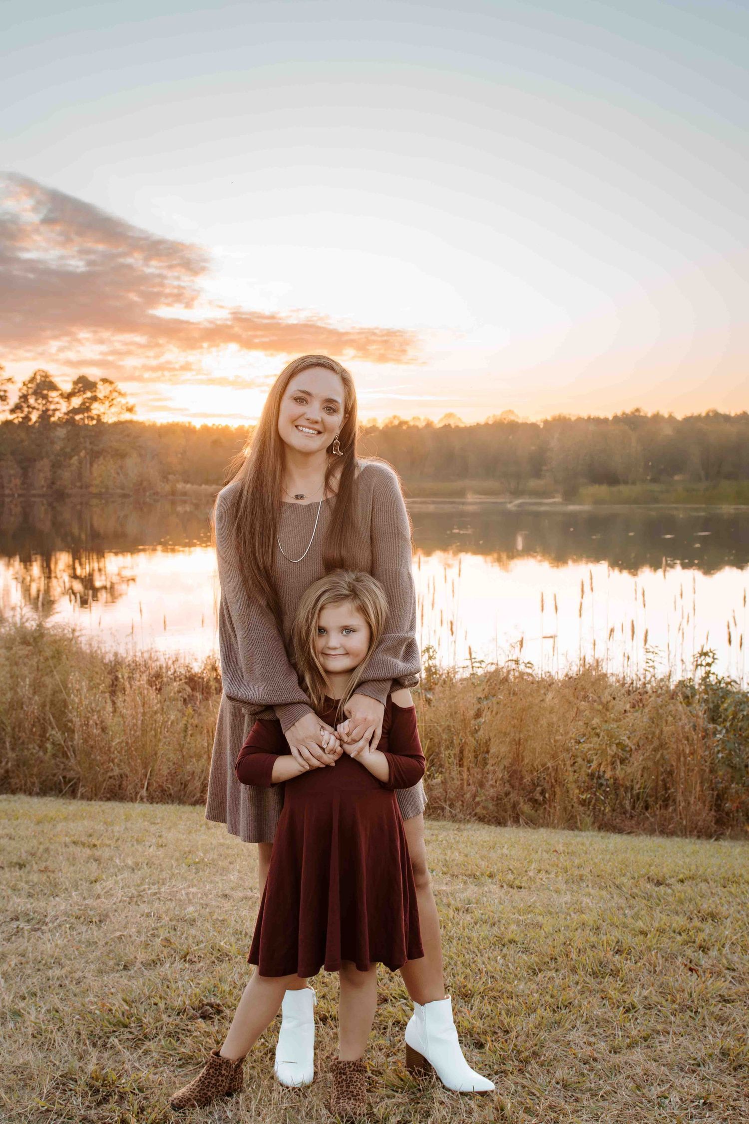Two people in dresses stand embracing by a lake at sunset with golden light reflecting off the water in the background.