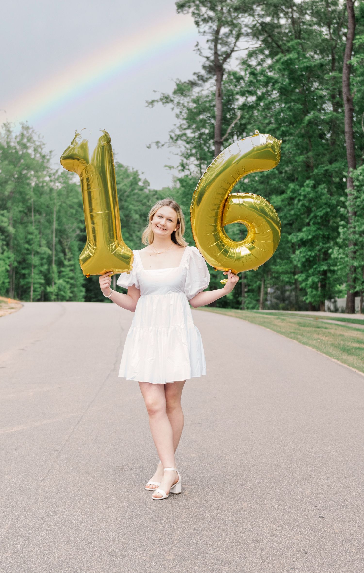 Young person in white dress poses on road with gold number 16 balloons while rainbow appears in background.