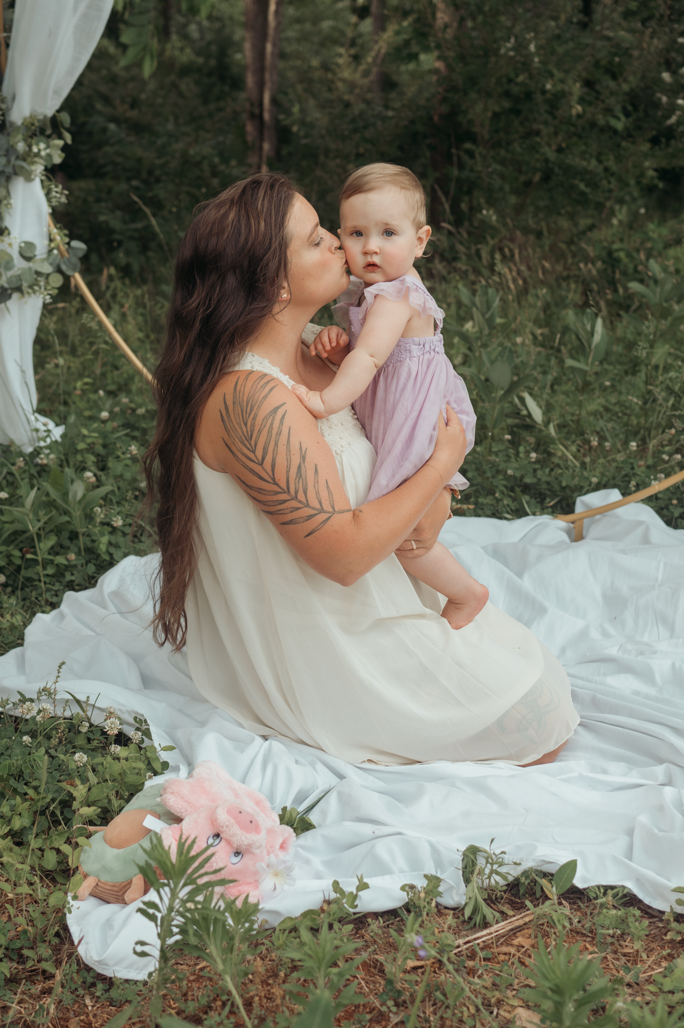 A heartwarming outdoor portrait of a parent and baby sitting on a white blanket with pink flowers nearby in nature.