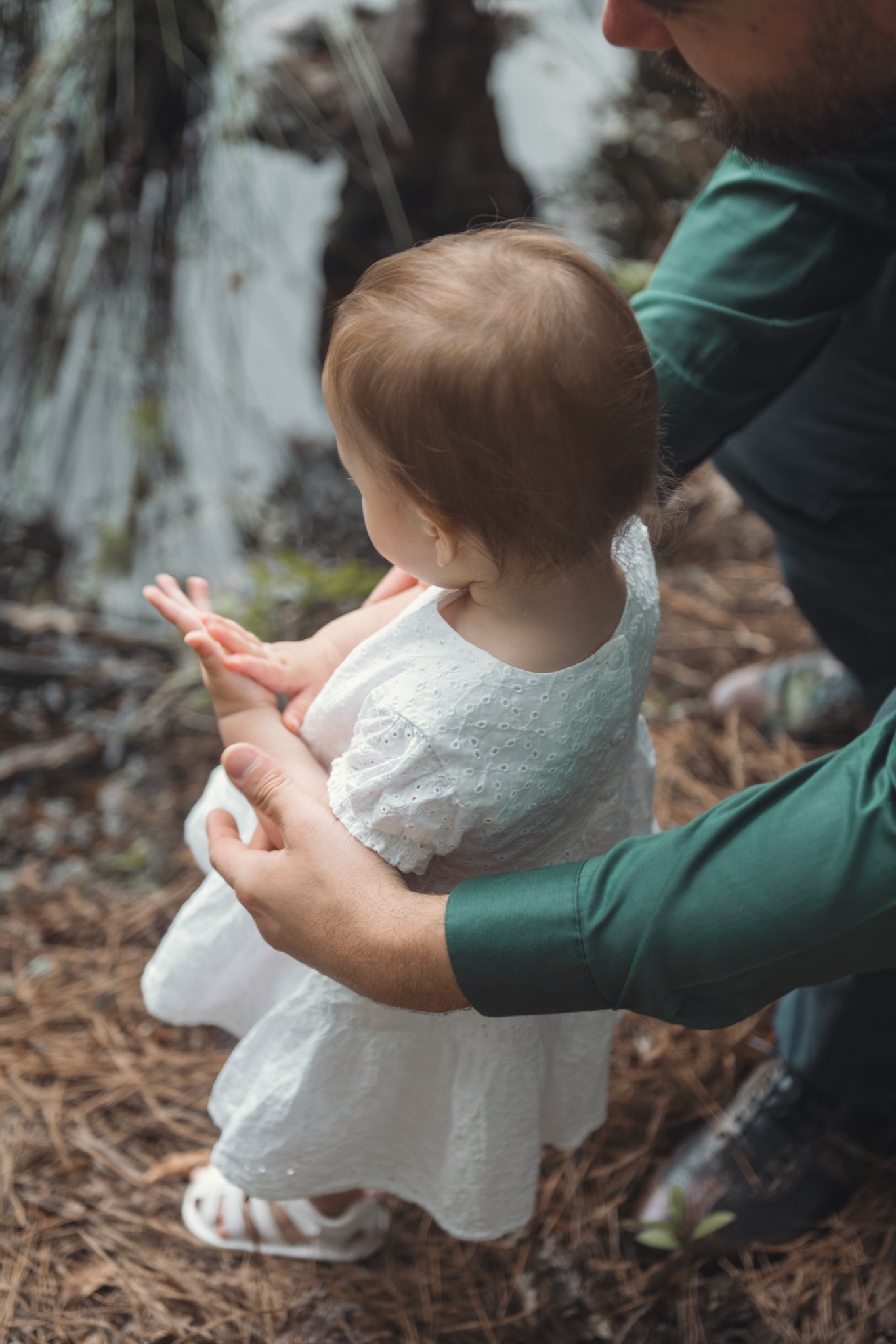 A baby in a white dress stands outdoors while being supported by someone wearing green.