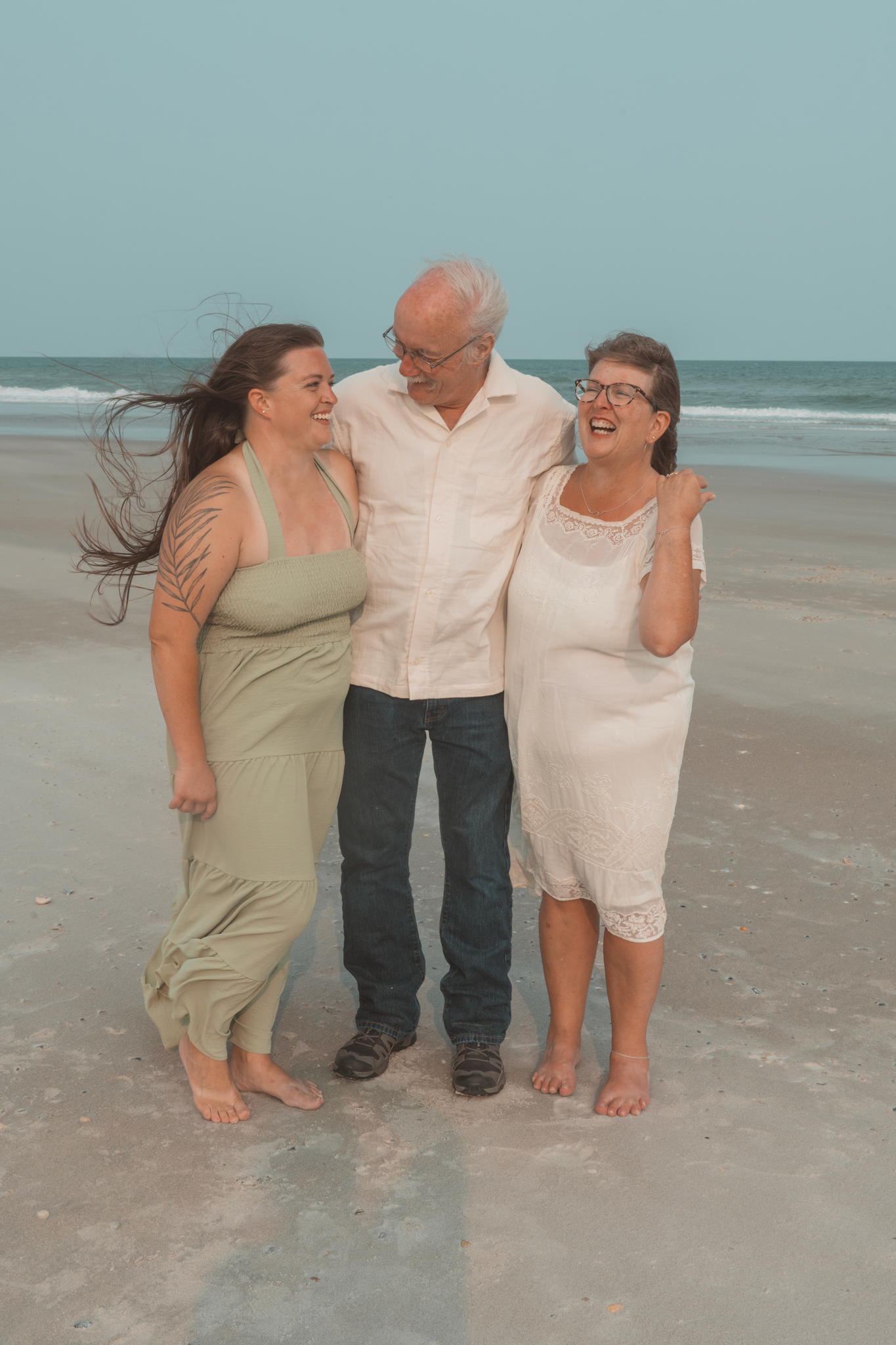 Three people share a joyful moment together while standing barefoot on a beach at dusk.