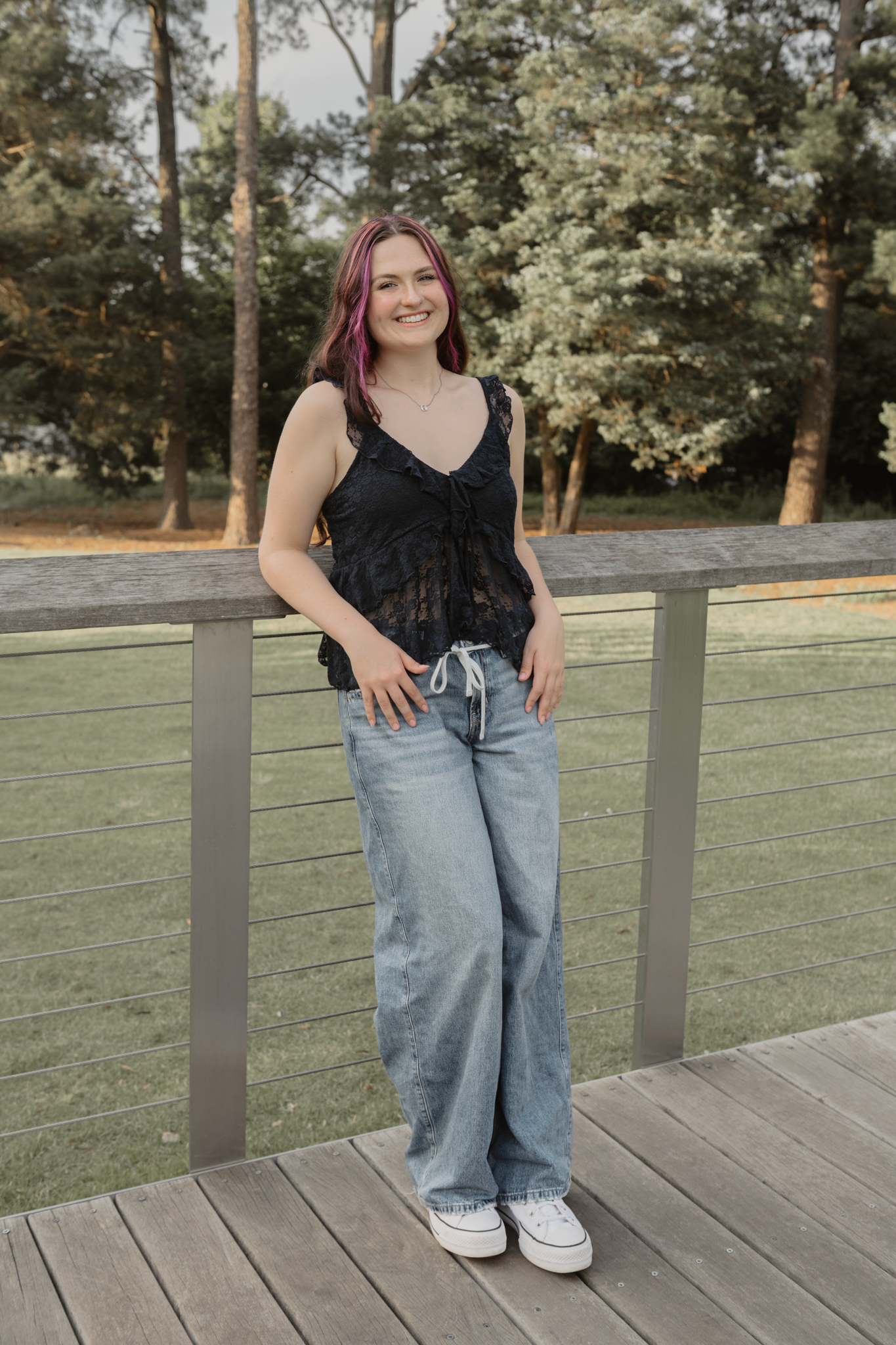 Haylee wearing a blue tank top and light wash baggy jeans stands on a wooden deck near flowering trees during her senior session