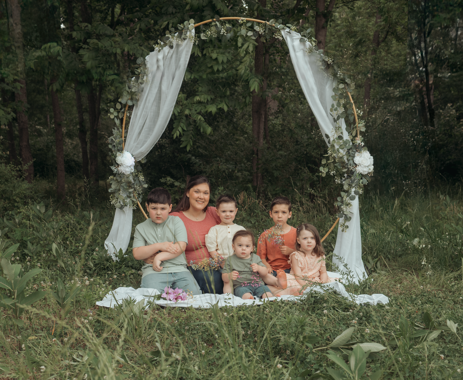 A group sits outdoors under an elegant white arch decorated with flowers and fabric in a natural woodland setting.
