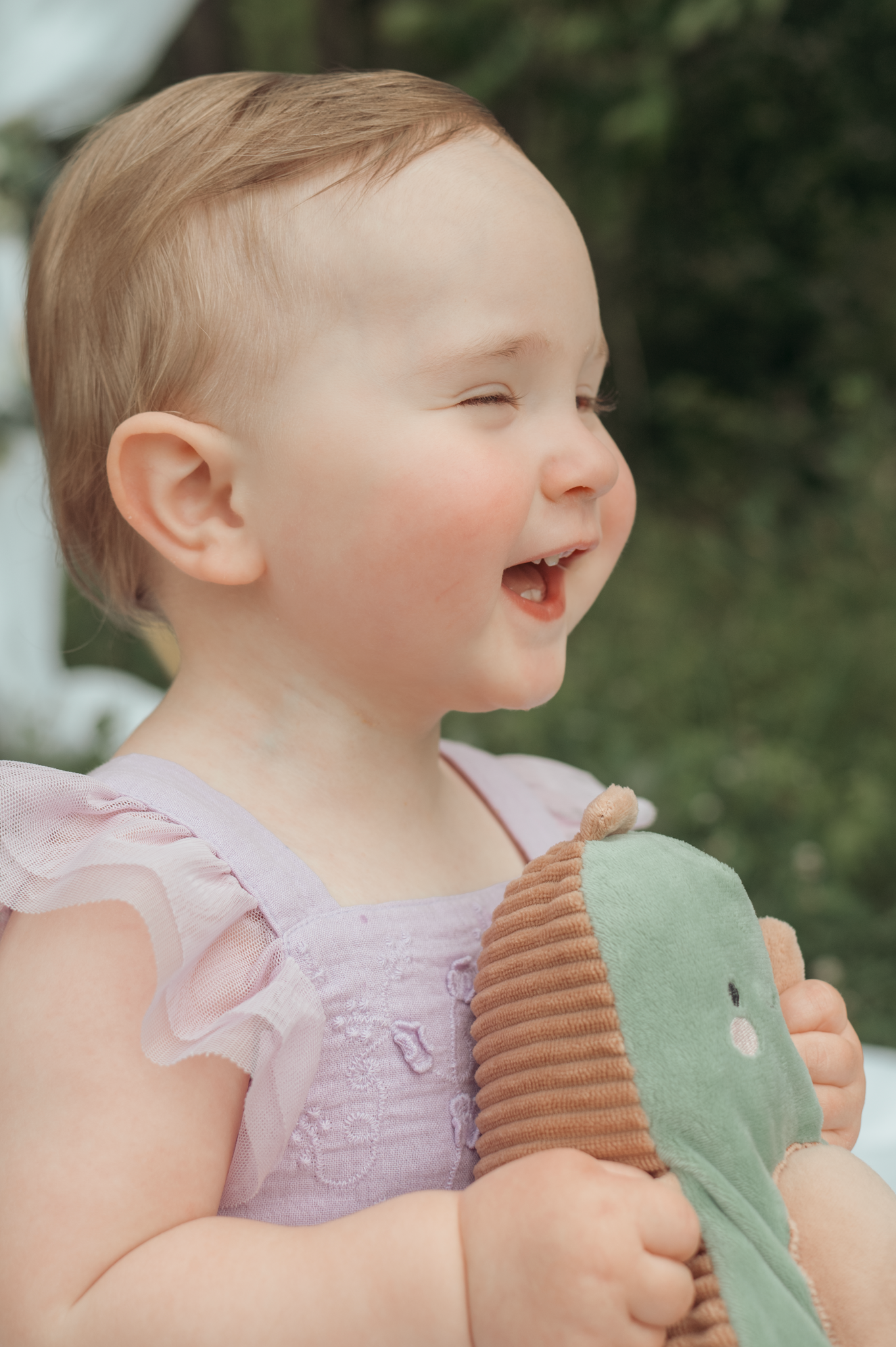 Joyful baby in pink dress laughing while holding a plush green toy bird outdoors.