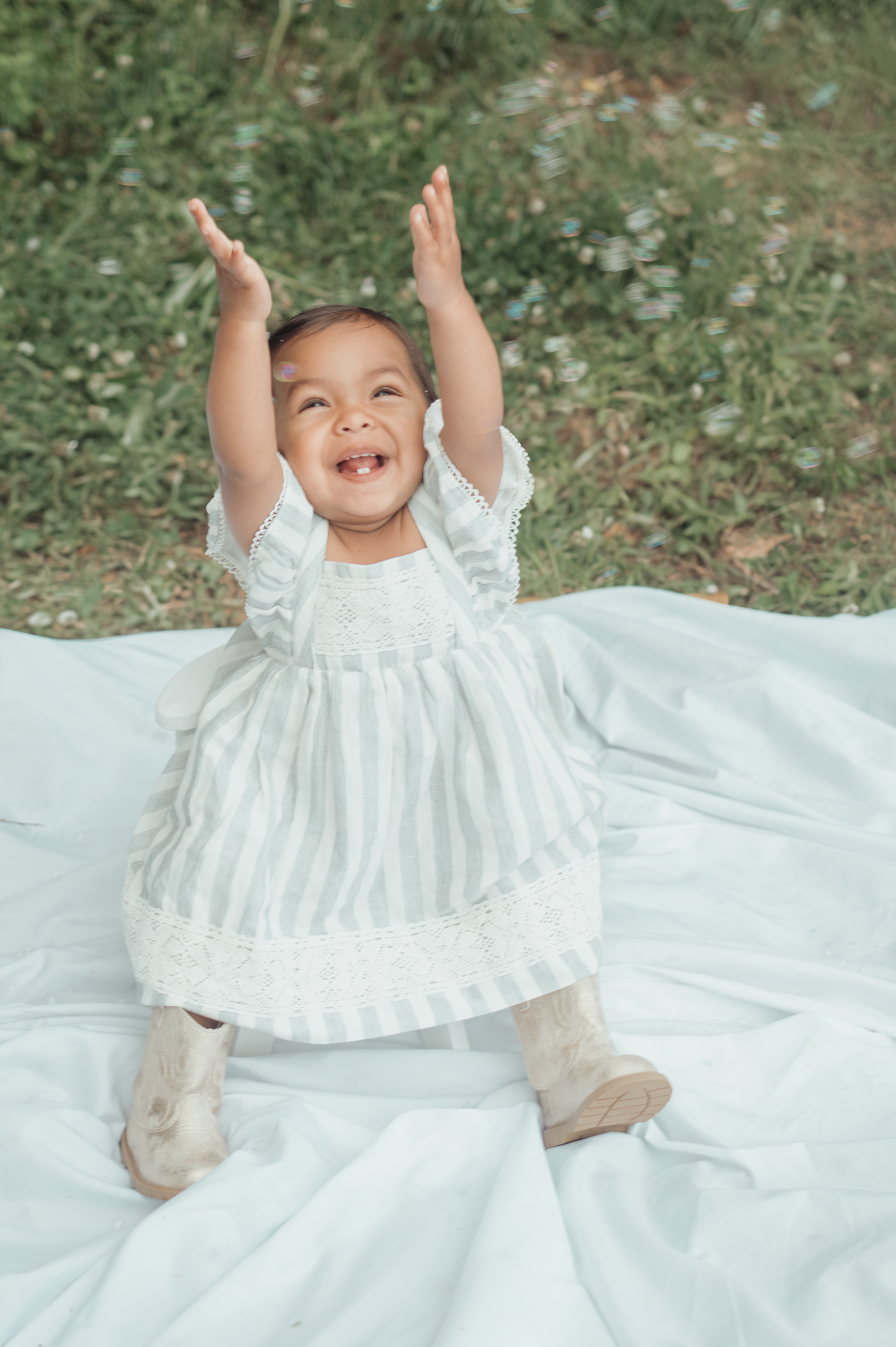 Joyful toddler wearing a white striped dress sits on a blanket outdoors with arms raised in celebration.