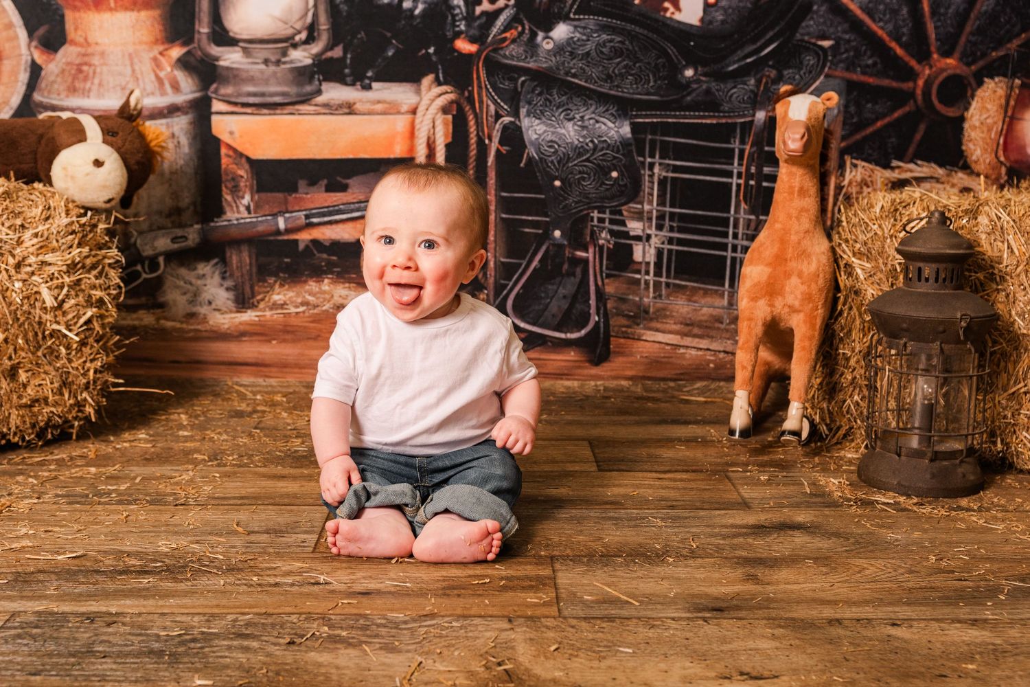 Baby sits on rustic wooden floor surrounded by hay bales, vintage suitcases and decorative props in studio setting.