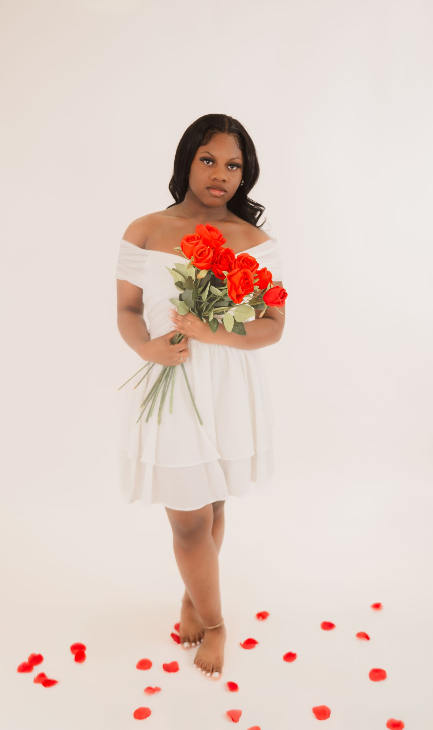 A barefoot woman in a white off-shoulder dress holds vibrant red roses while standing on scattered rose petals.
