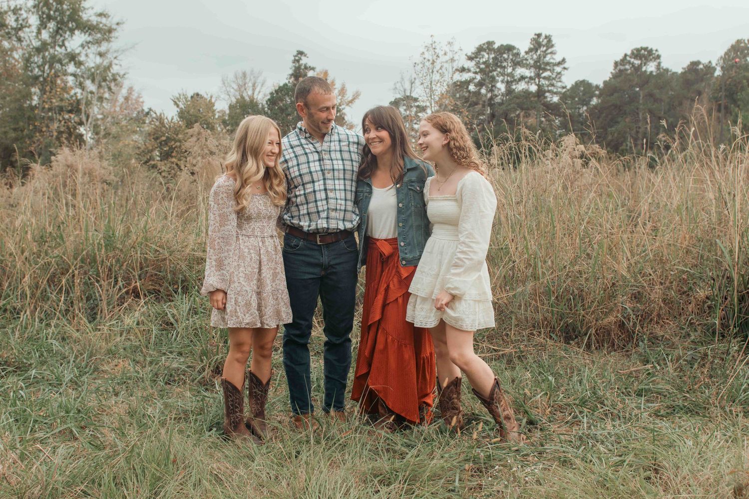 A group of friends in boho-style dresses and boots stand together in a golden autumn field at sunset.