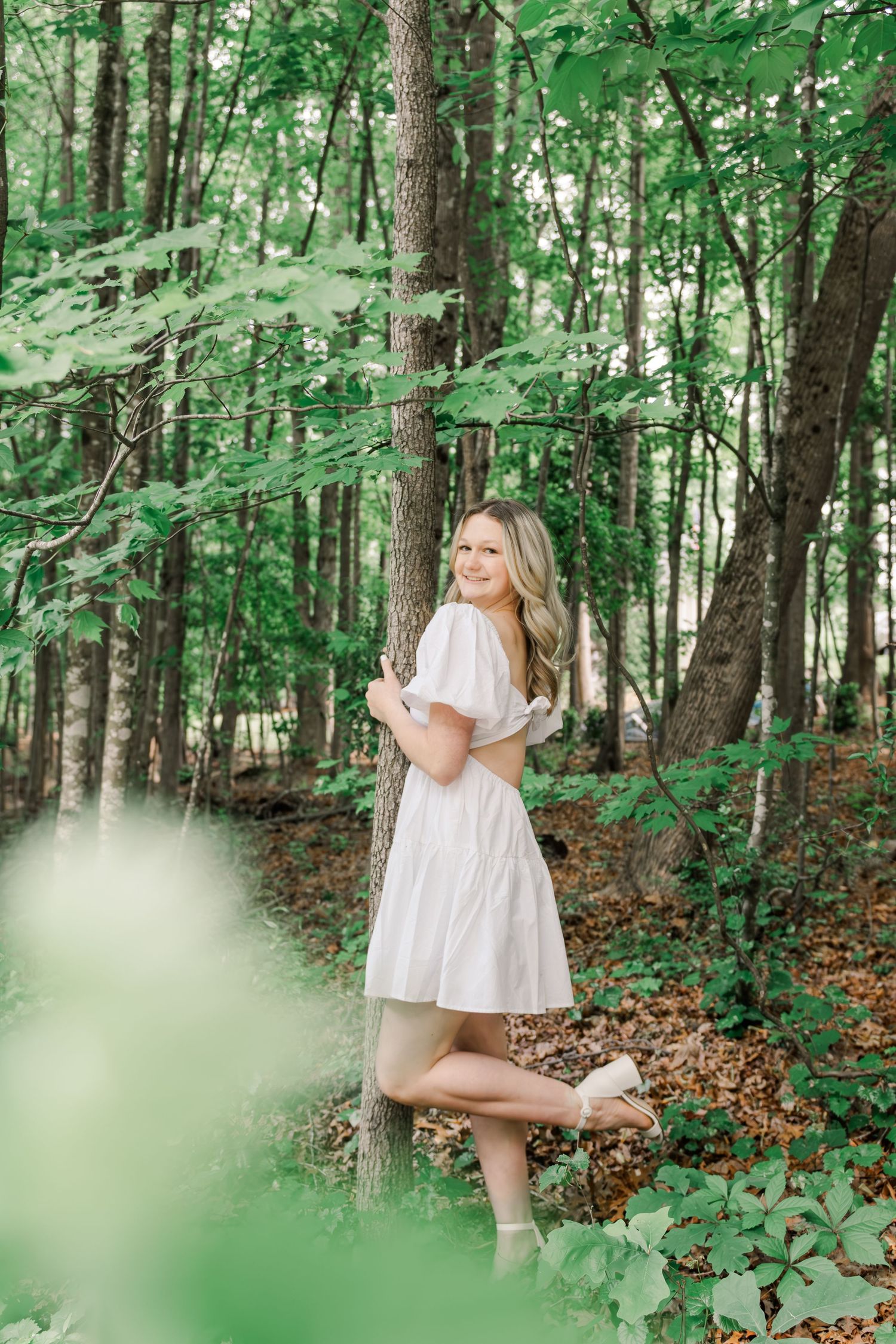 A white sundress stands out against the lush green forest backdrop as light filters through summer foliage.