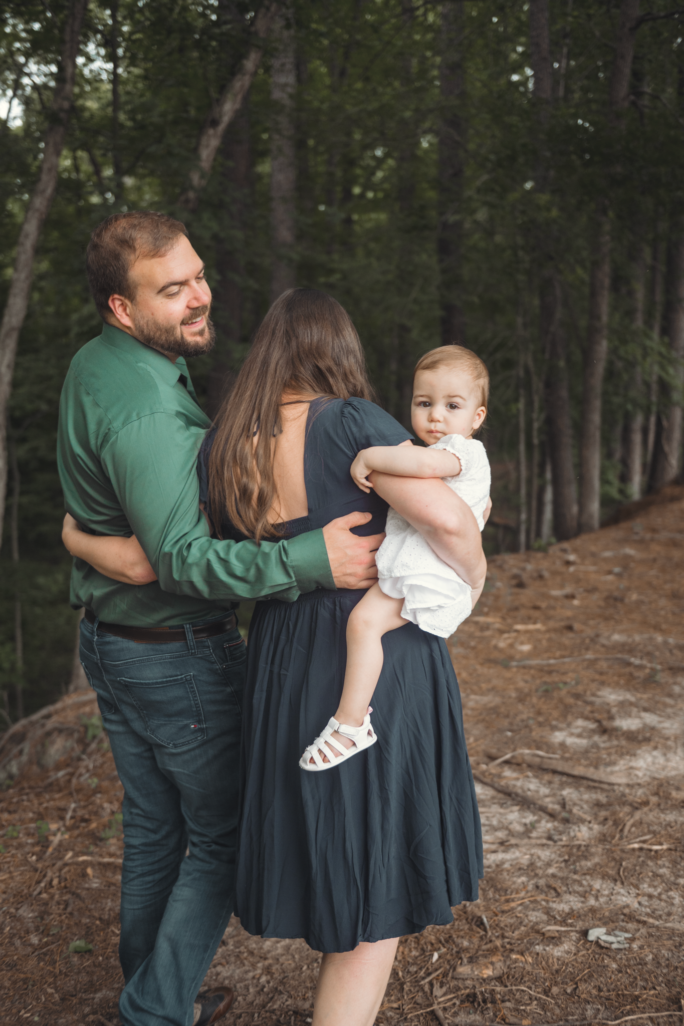 Young family shares tender moment together on wooded trail with towering trees in background.