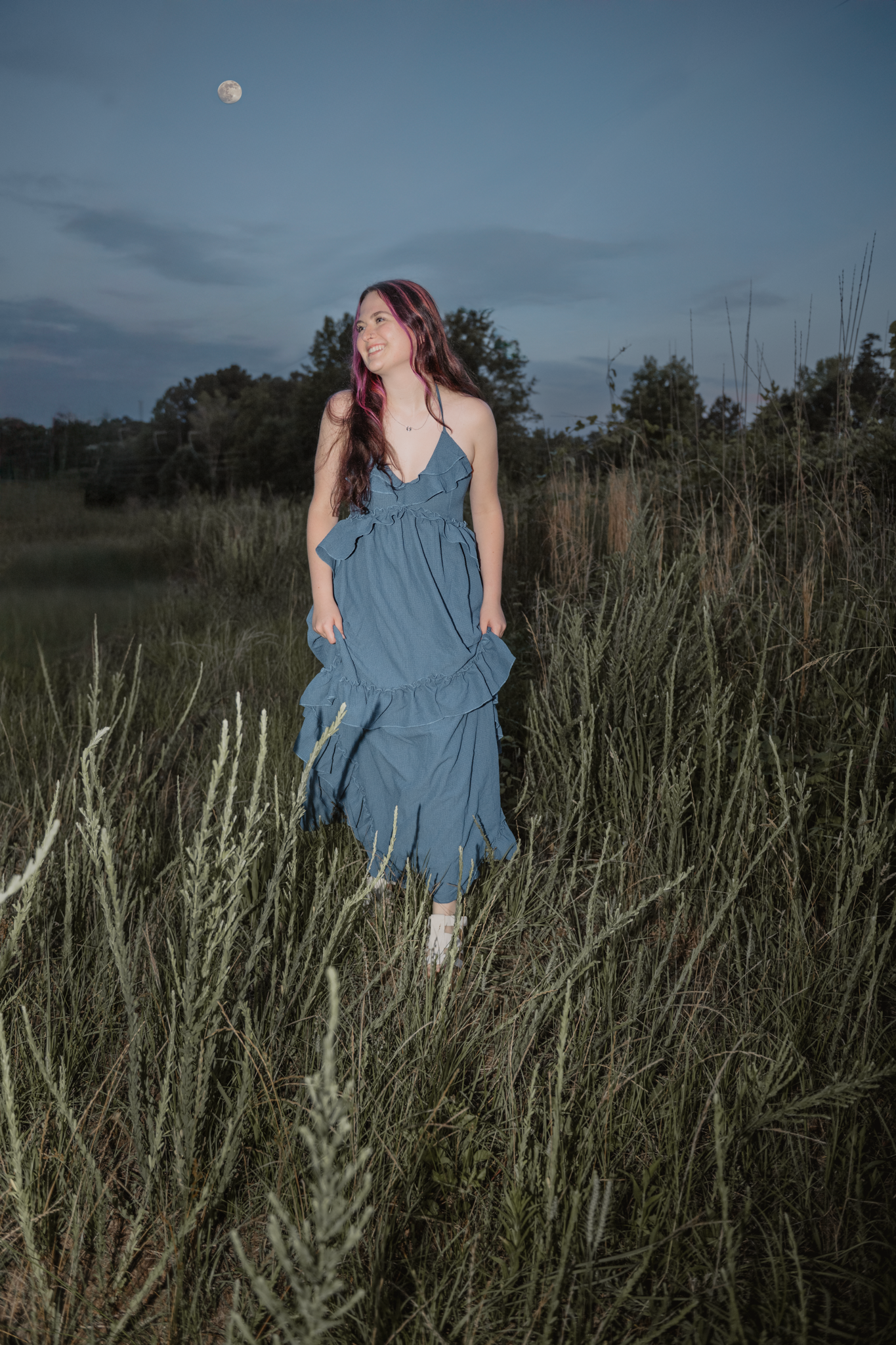 Person in a blue dress stands thoughtfully among tall grasses and reeds at dusk.