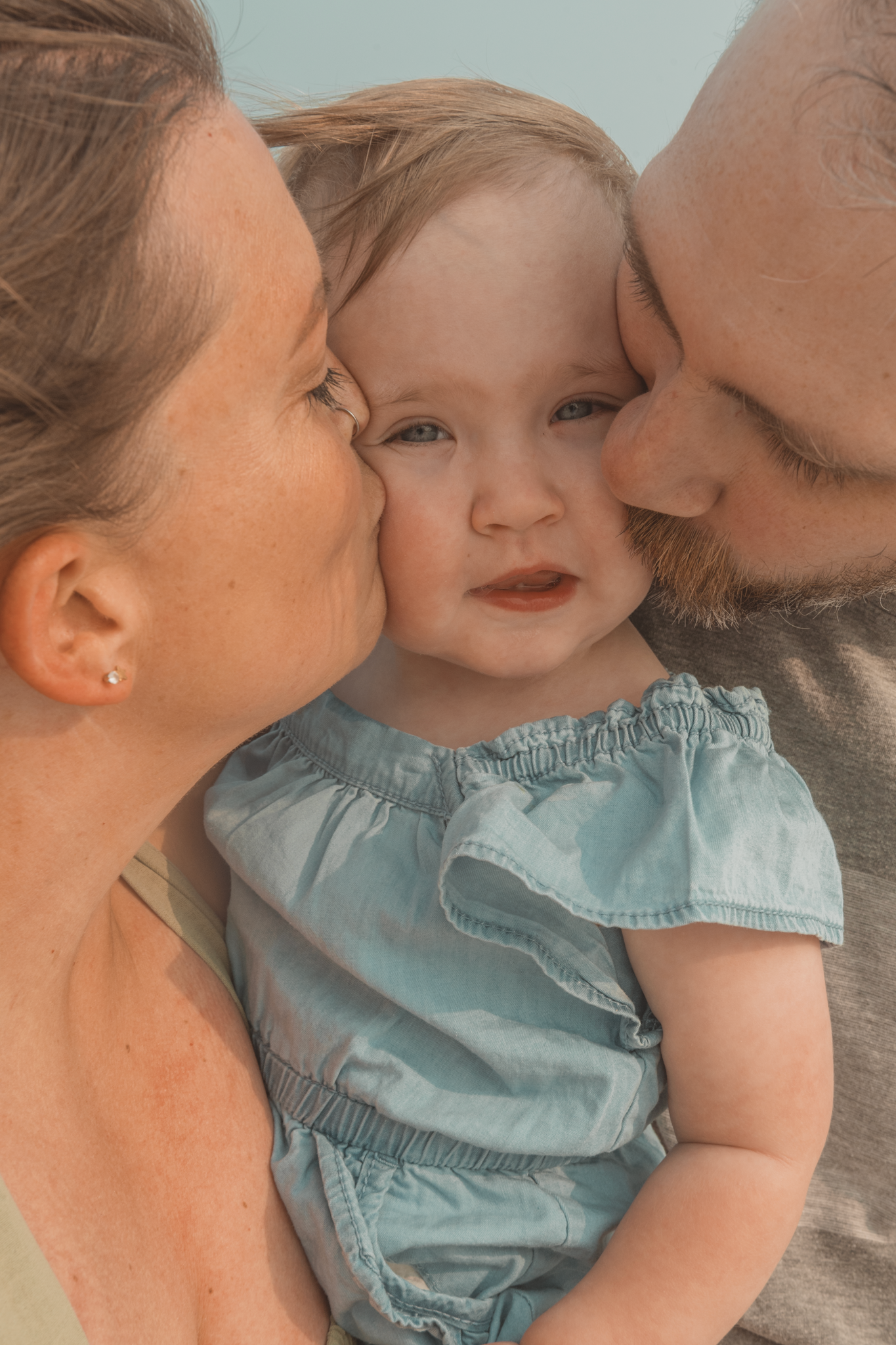 A close-up tender moment of parents kissing their baby wearing a light blue outfit.