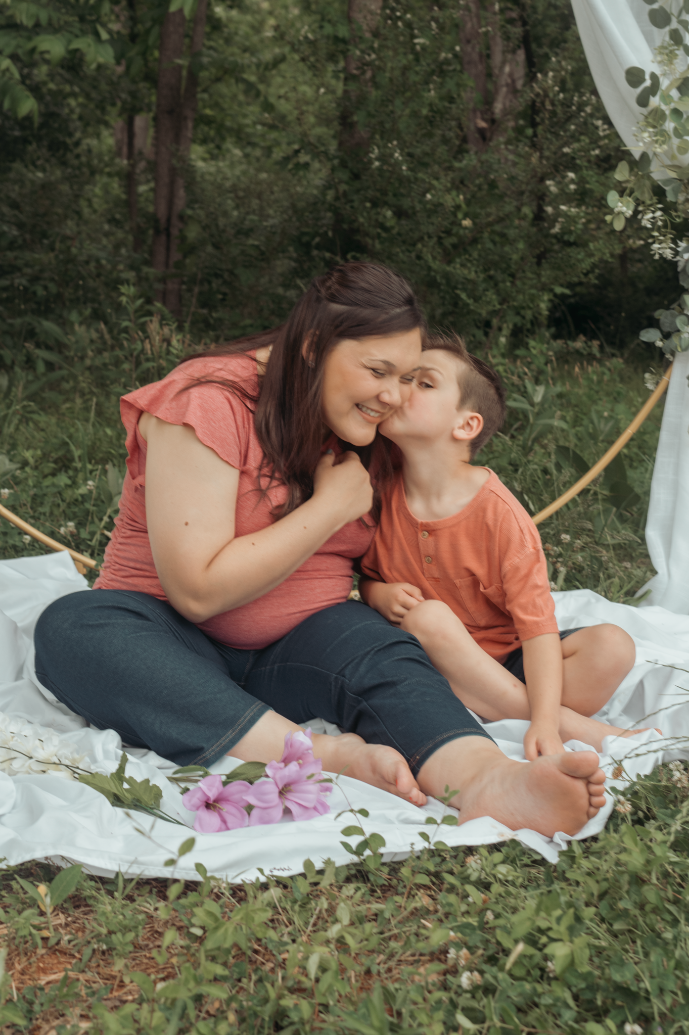 A tender outdoor portrait showing affection on a white blanket with pink flowers in a wooded garden setting.