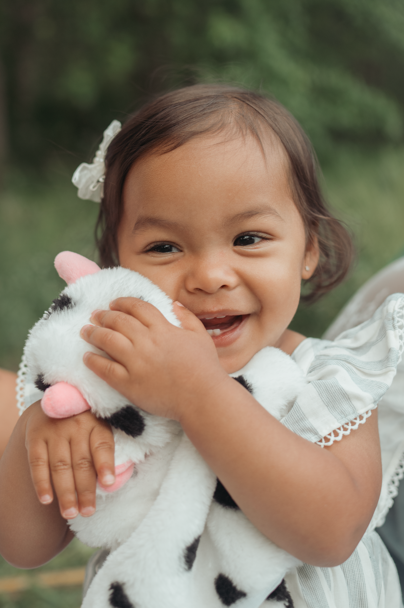 Toddler in white dress laughs joyfully while hugging a plush white and black spotted toy animal.