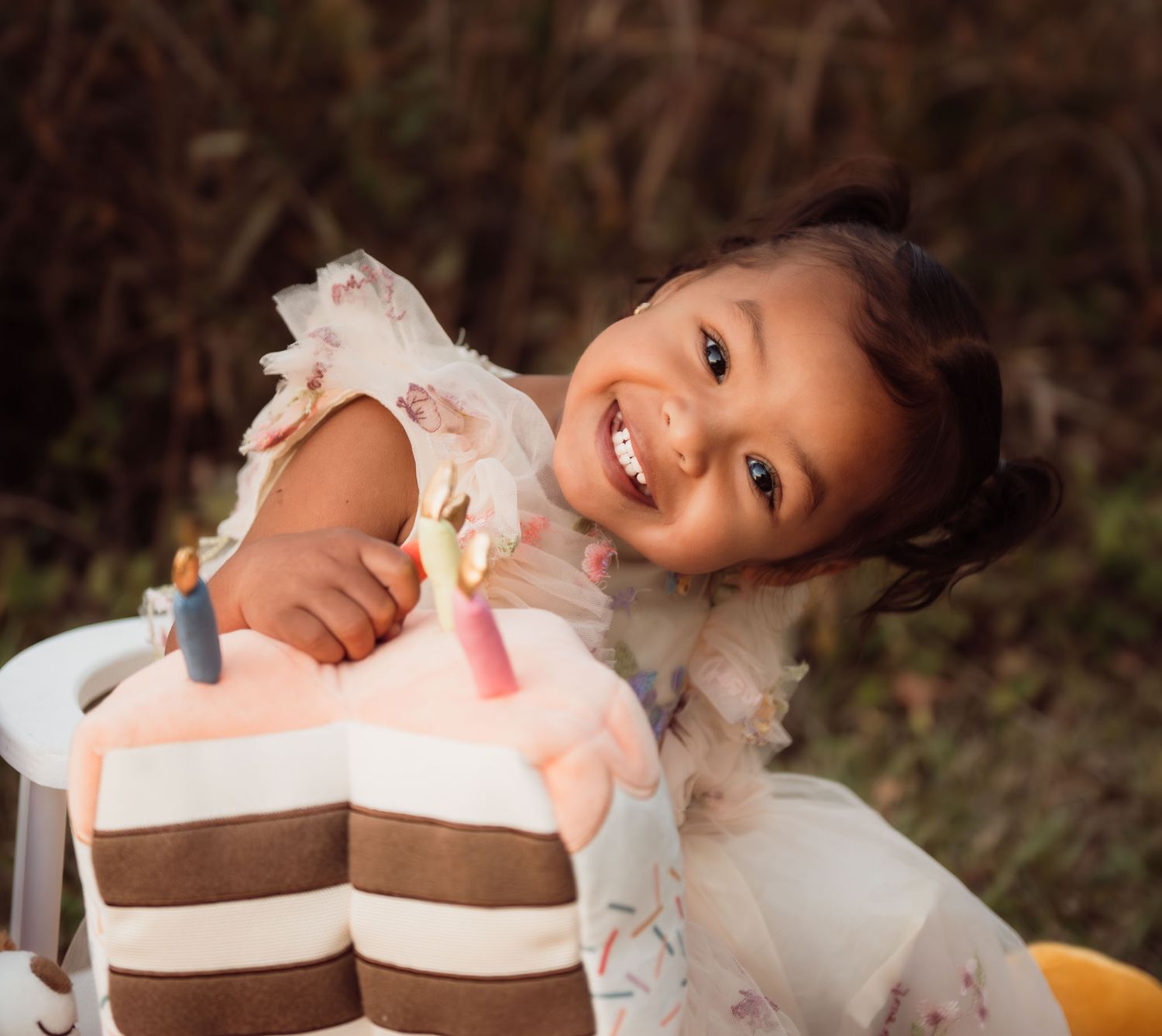 A smiling child in a white dress leans over a birthday cake with pink and brown frosting.
