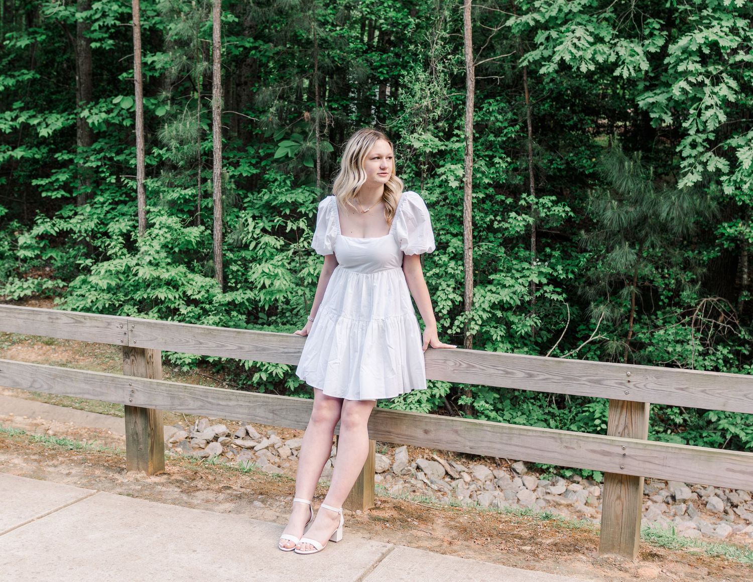 A person in a white summer dress and pink shoes poses casually along a wooden railing in a wooded area.