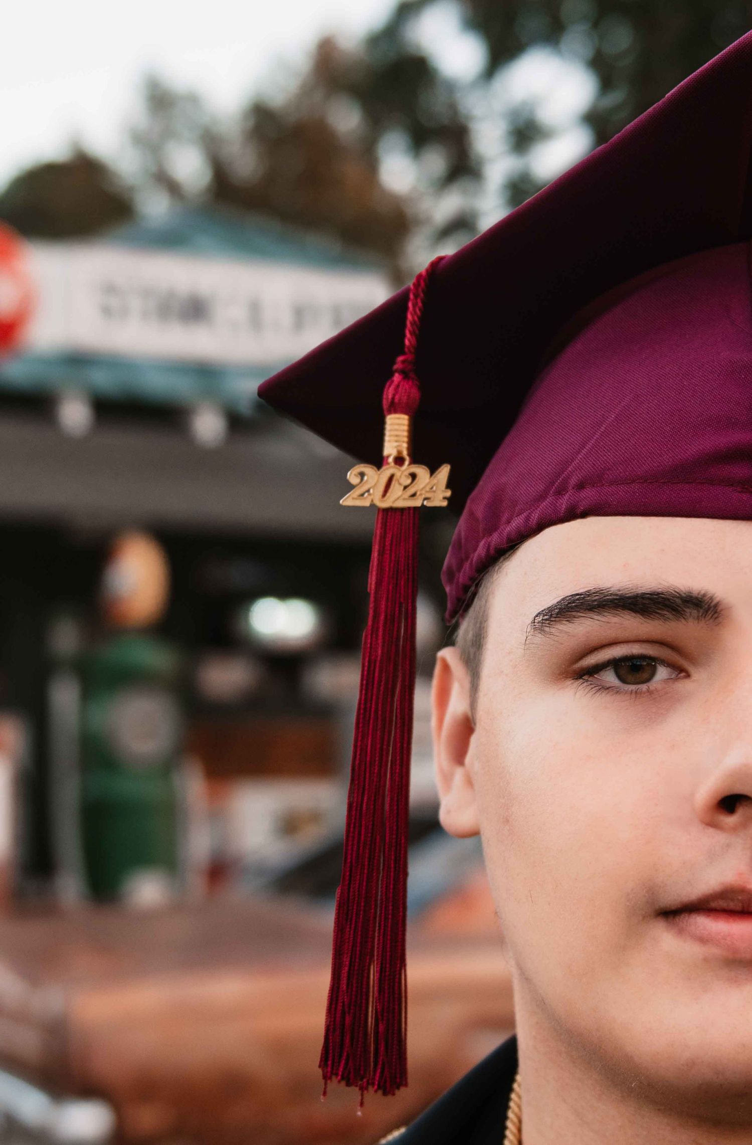A close-up graduation portrait showing a maroon cap with a 2024 tassel against a blurred outdoor background.