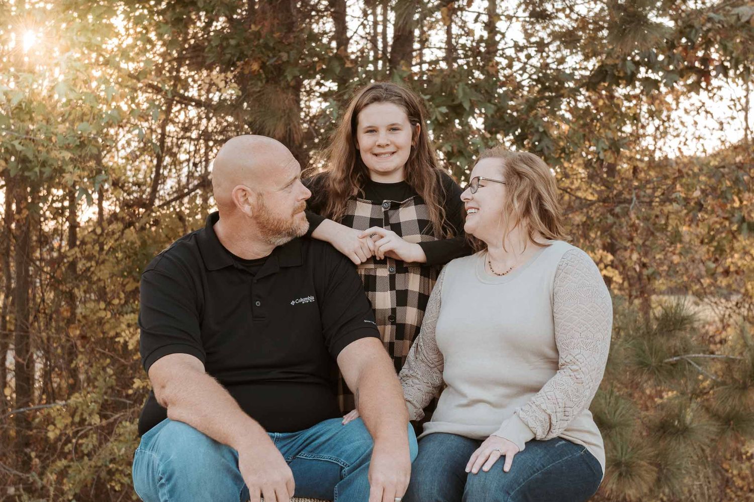 Outdoor autumn family photo with warm sunlight filtering through trees in a natural woodland setting.