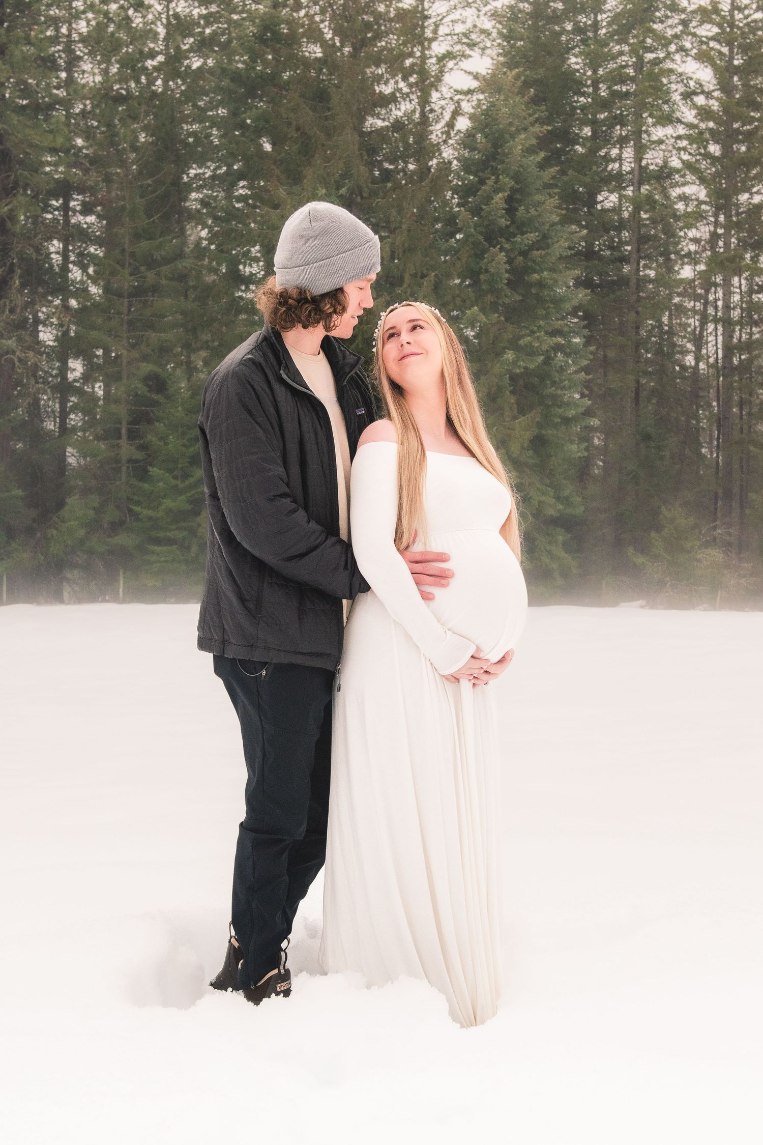 A romantic winter engagement photo showing a couple in white and black standing together in a snowy forest setting.