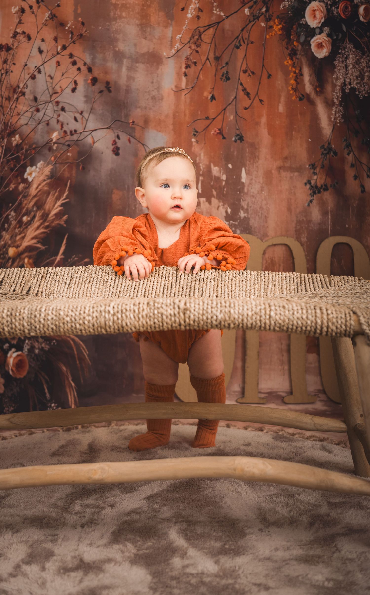 A small child in an orange outfit stands at a rustic woven bench against an autumn-themed studio backdrop.