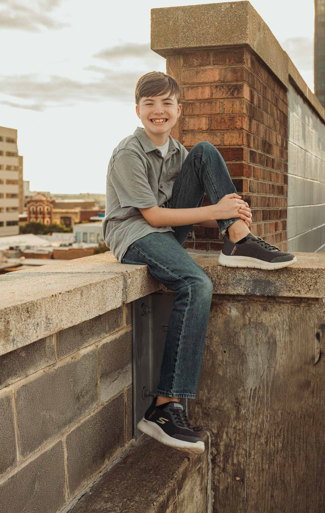 A child in jeans and sneakers sits on a concrete ledge at sunset against an urban brick building backdrop.