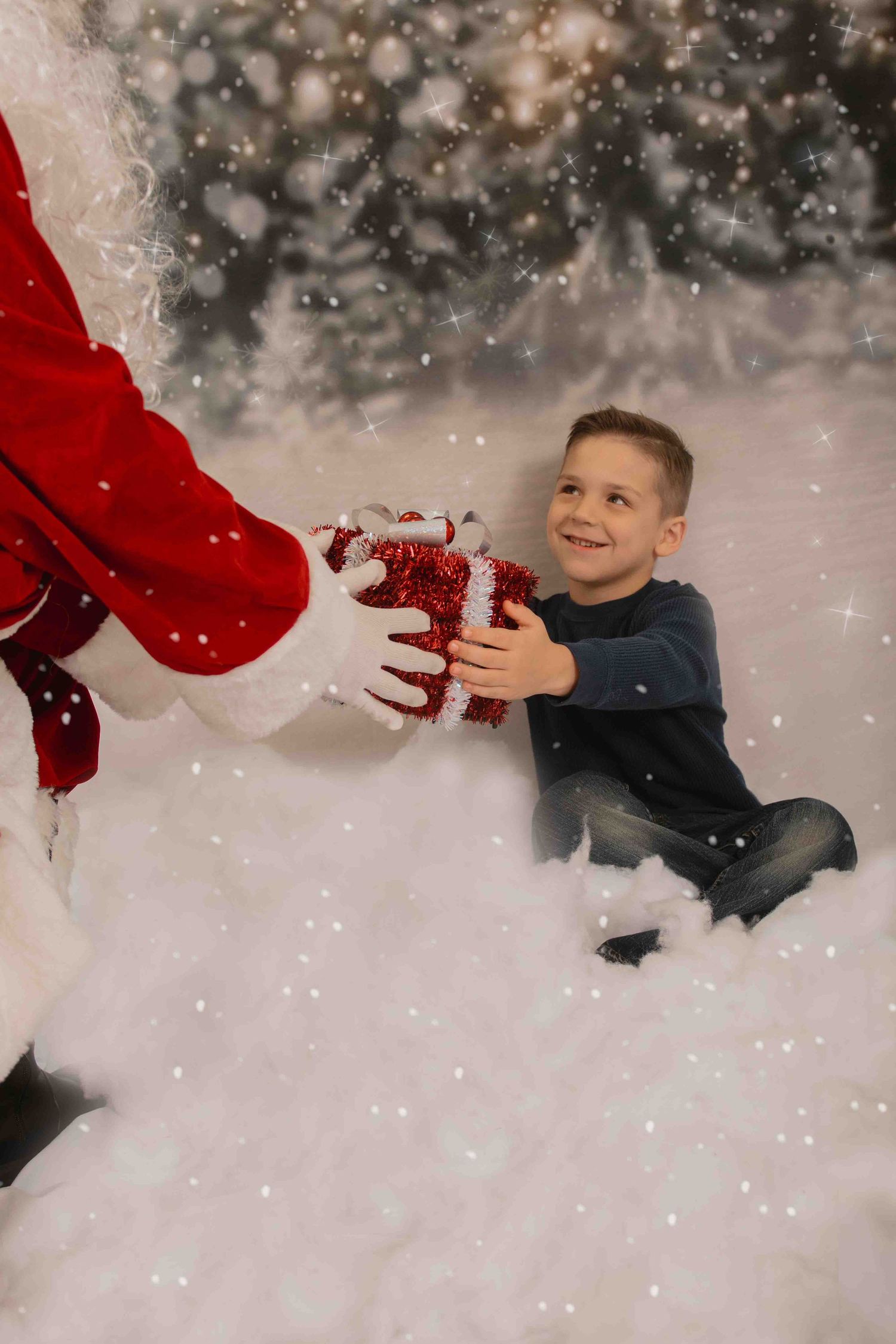 A young child receiving a gift from someone in a red Santa suit while sitting on snowy ground with sparkles in the background.