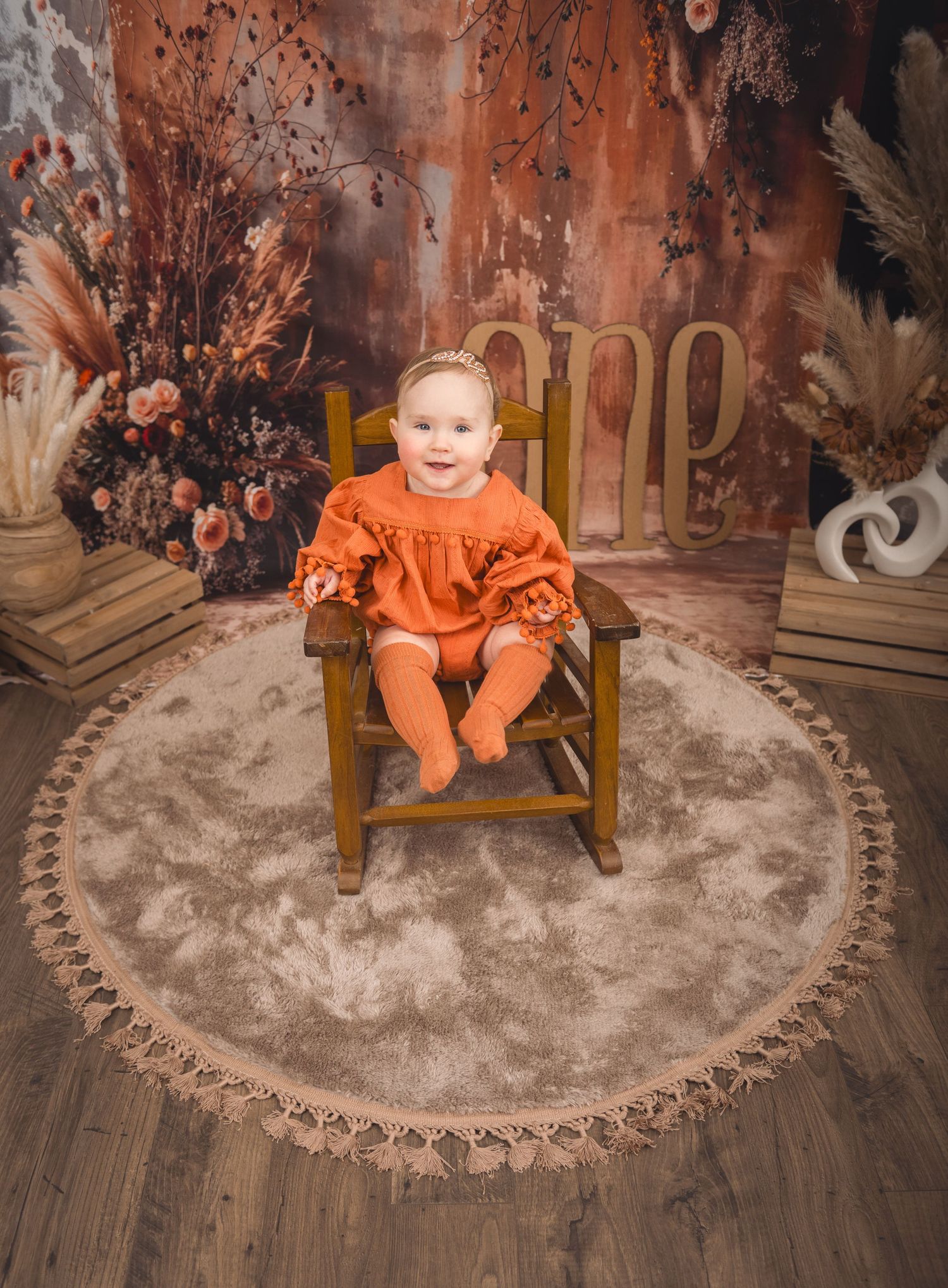 A small figure in a bright orange outfit sits in a wooden chair on a round rug surrounded by autumn-themed decor.