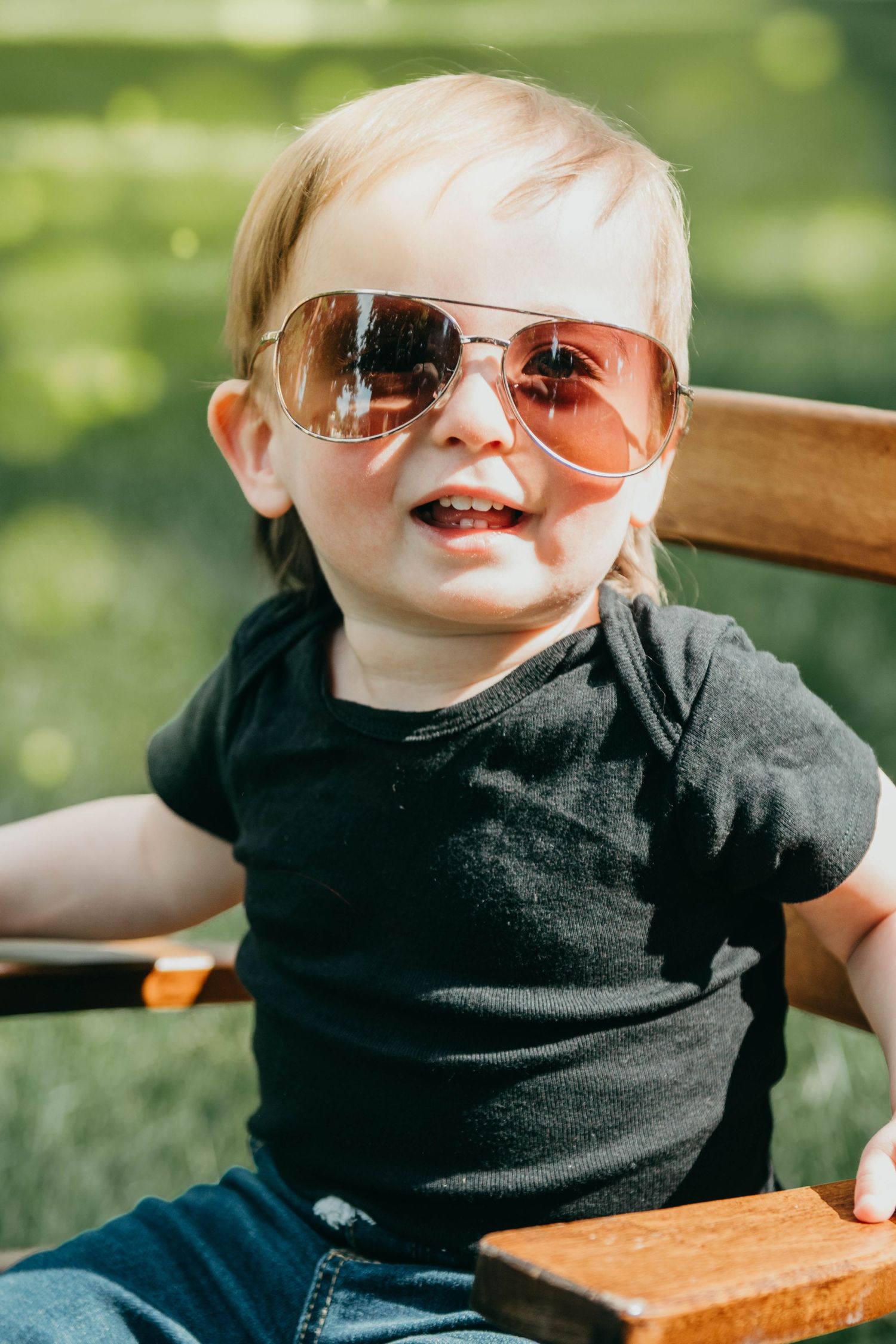 Child wearing aviator sunglasses and black t-shirt sits on wooden chair while smiling on sunny day.