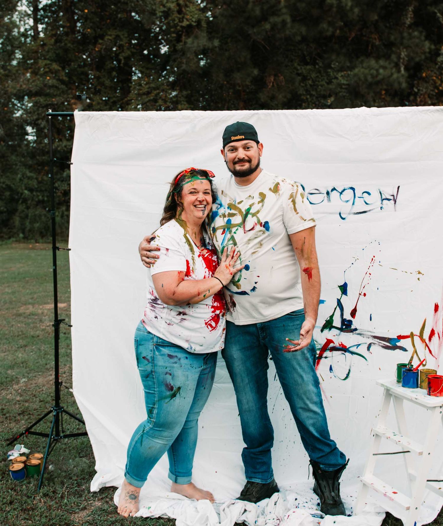 Paint-splattered couple poses in front of white backdrop during outdoor art session.
