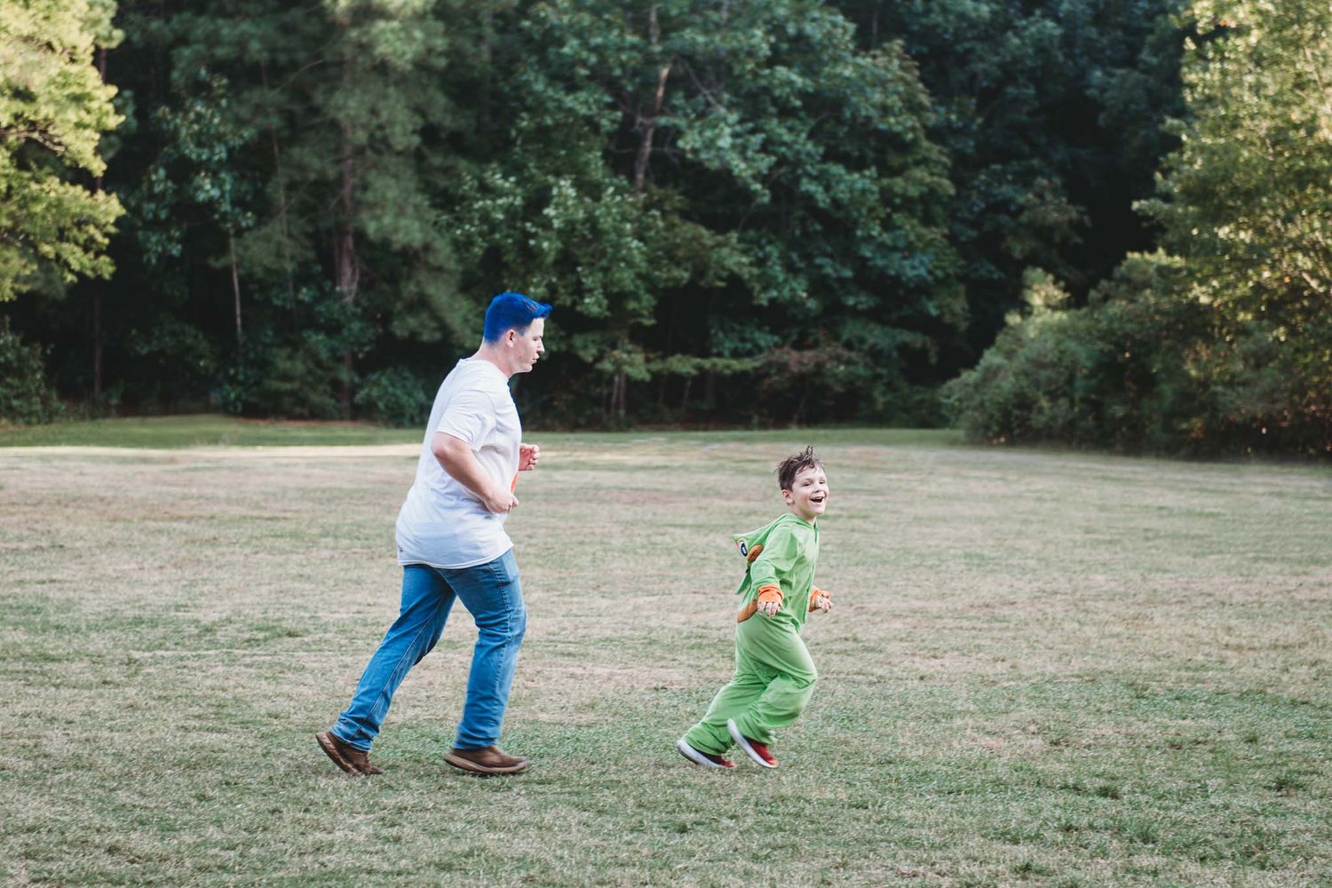 A parent and child wearing bright clothing play together in a grassy field surrounded by trees.