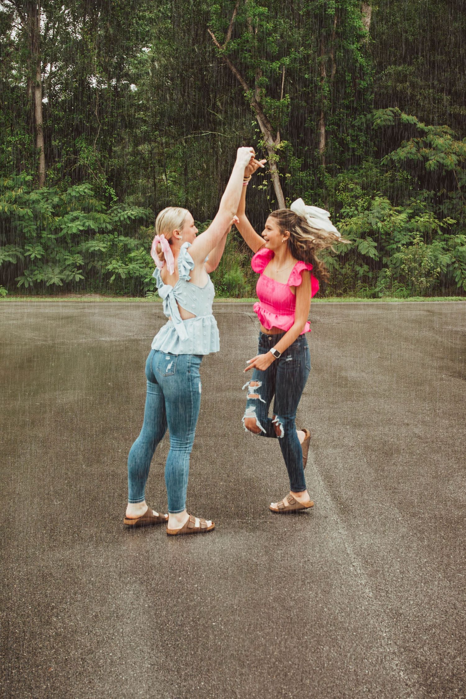 Two friends dance joyfully together on a wet asphalt surface with lush green forest in the background.