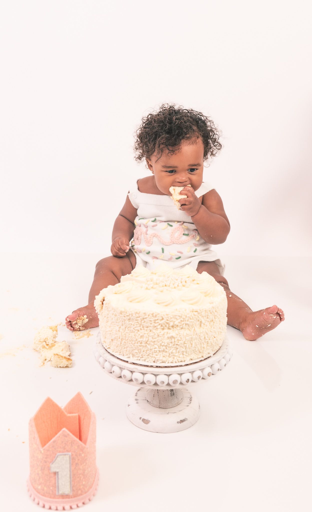A one year old in white outfit sits on a cake stand with a smashed birthday cake and coral crown nearby.