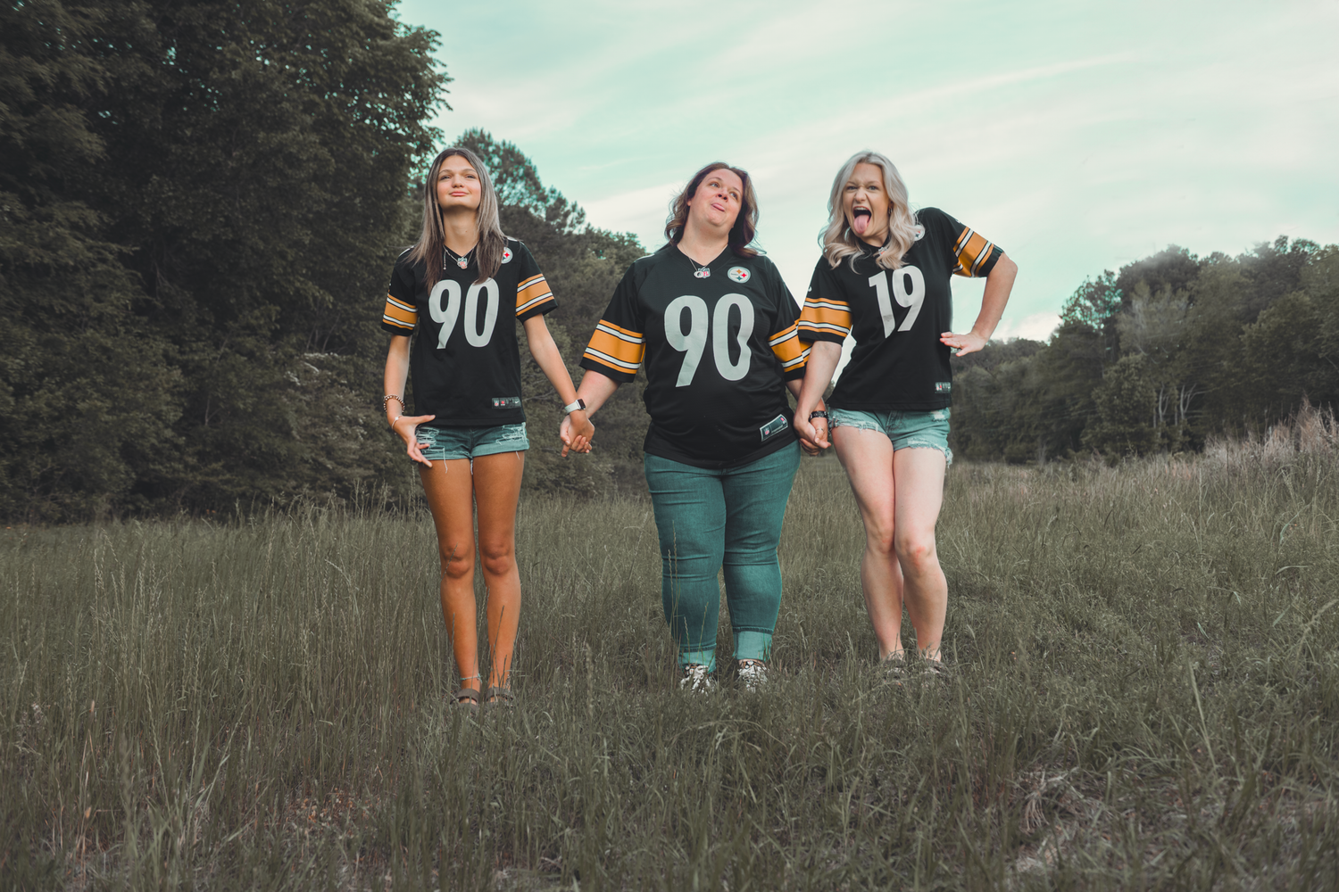 Three friends wearing matching black sports jerseys with number 90 and 19 pose together in a grassy field at dusk.