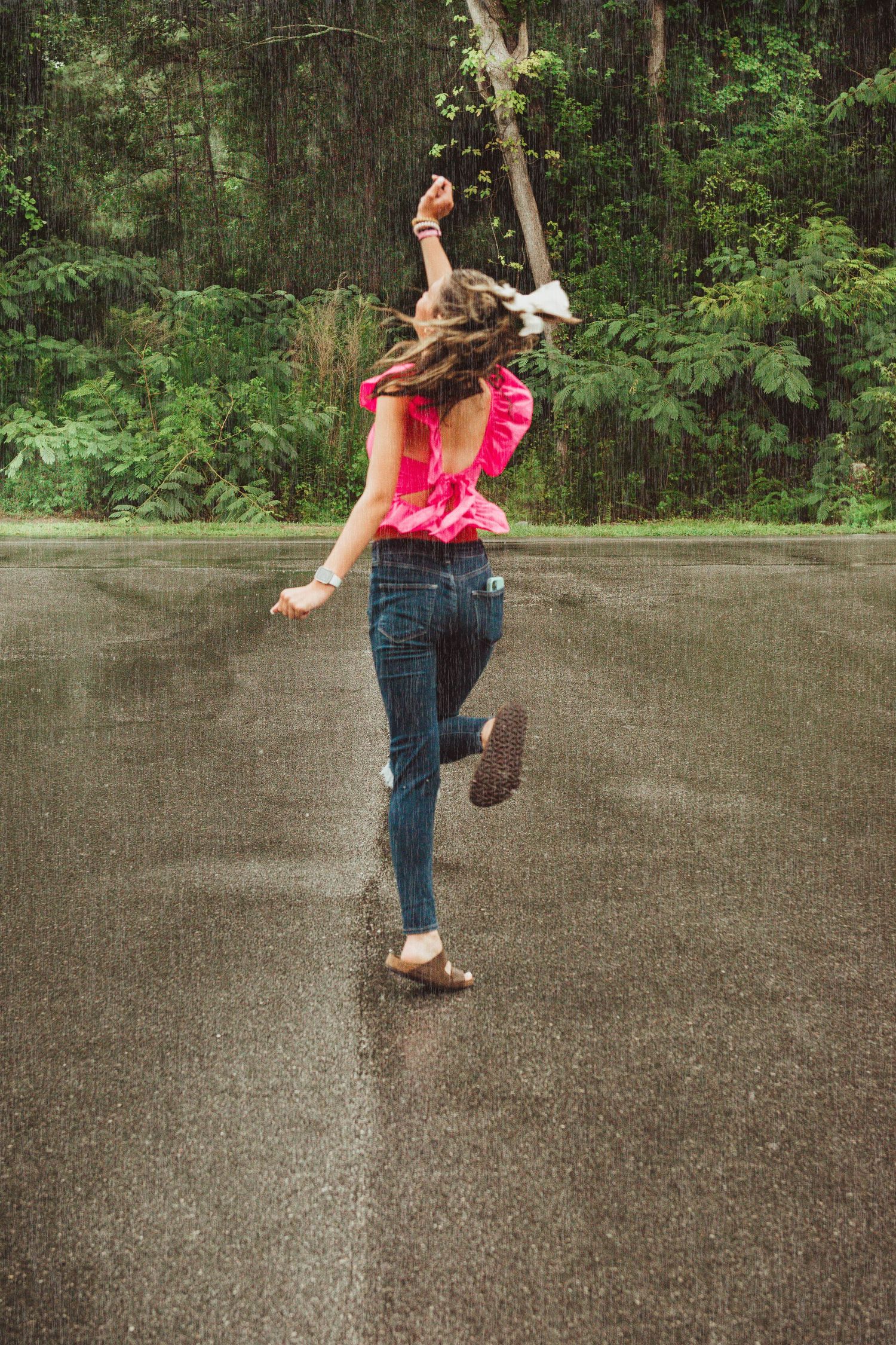 Someone in jeans and a pink top jumps joyfully on a wet pavement with green forest in the background.