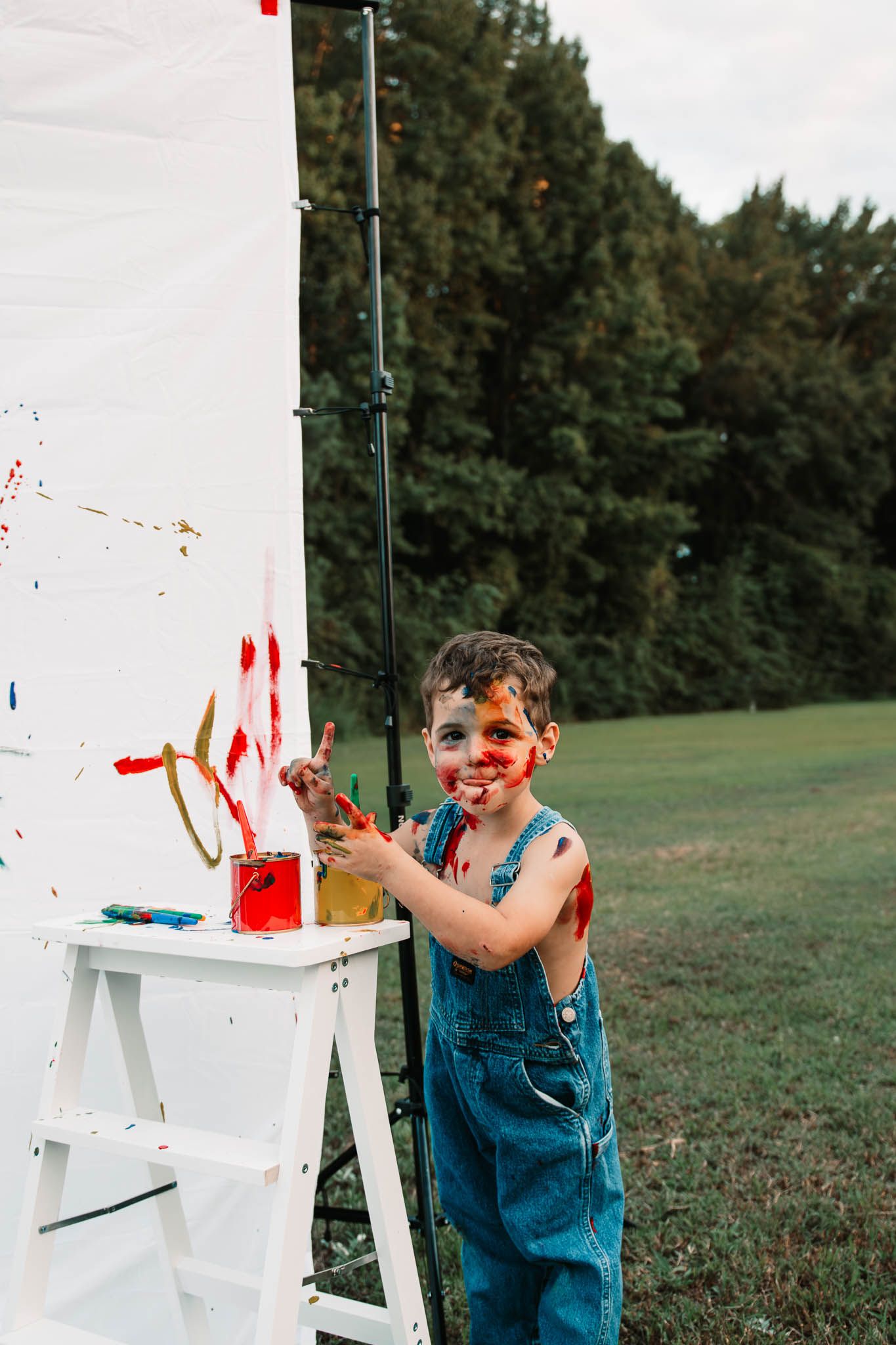 Smiling child in denim overalls having fun with paint and a white ladder outdoors in summer.