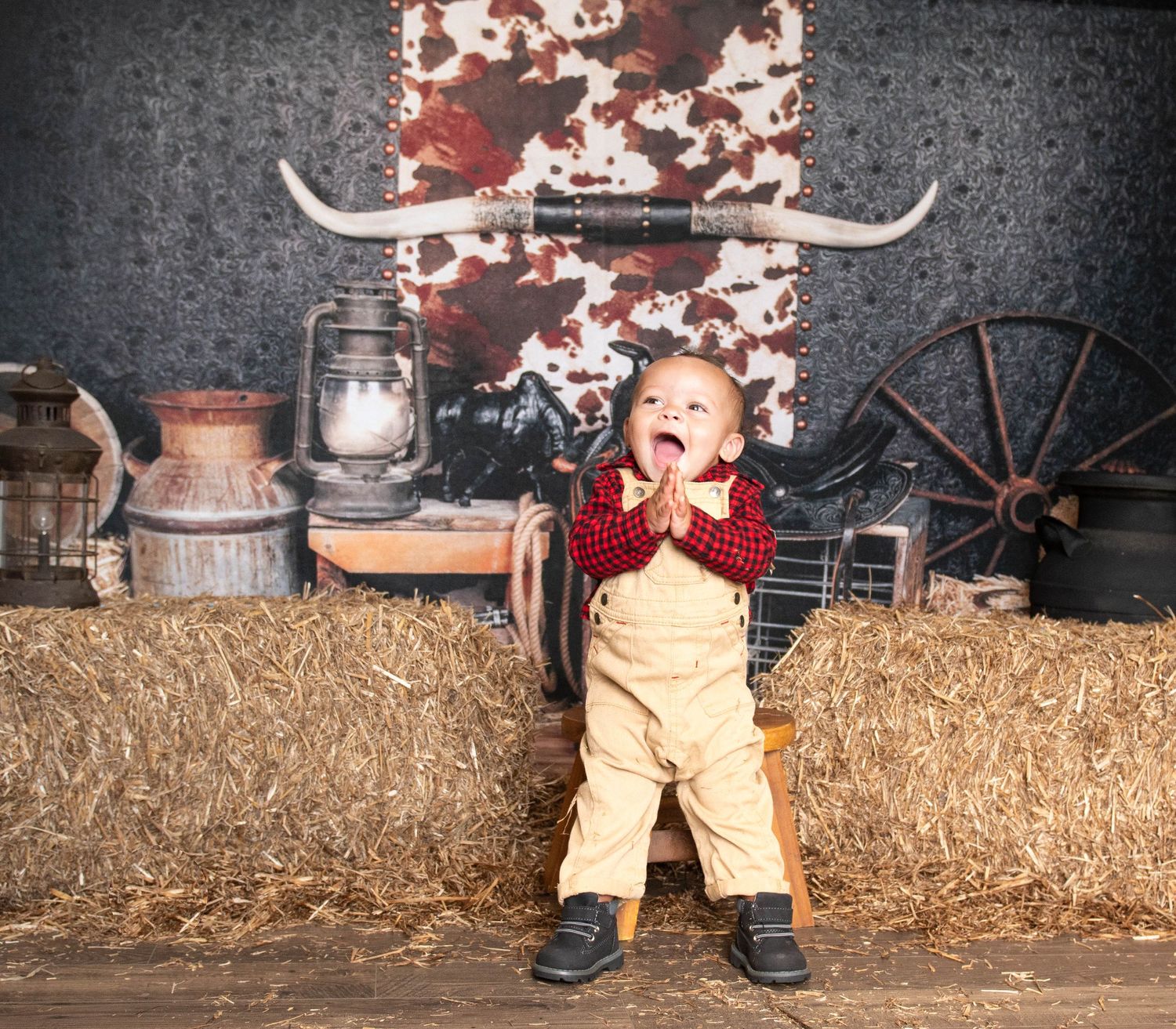 A small child in overalls and red top stands excitedly on hay bales against a rustic backdrop with vintage farm props.
