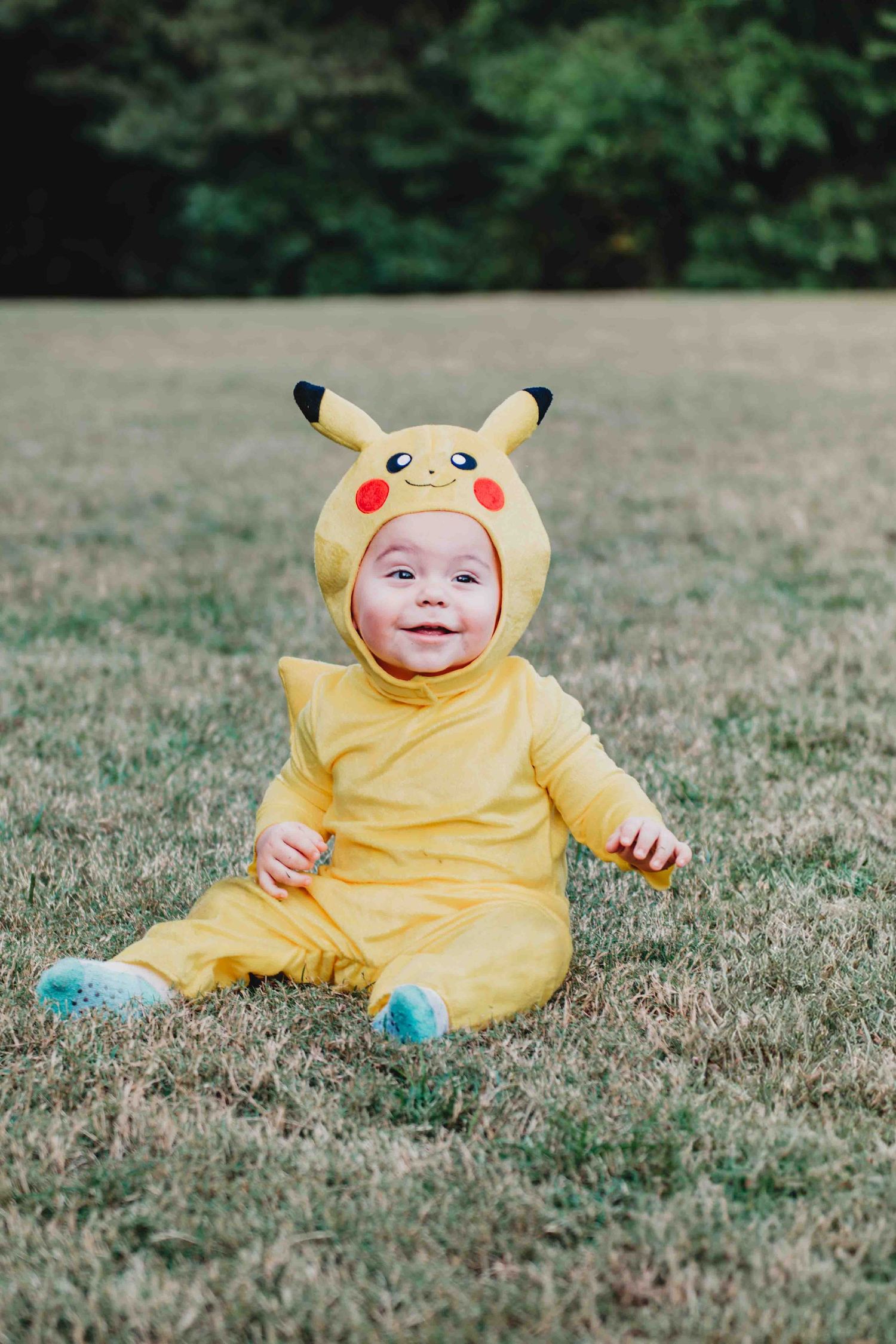 A small child wearing a yellow Pikachu costume sits on grass outdoors during a photo session.
