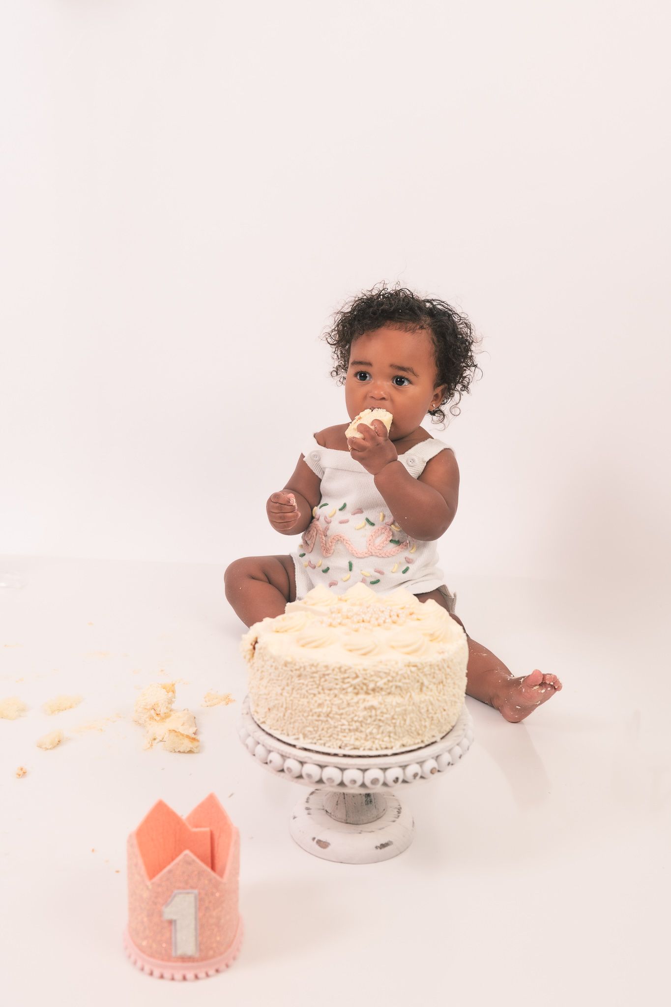 Small child in white outfit enjoying cake smash photo session with white frosted cake and coral crown prop on pink background.