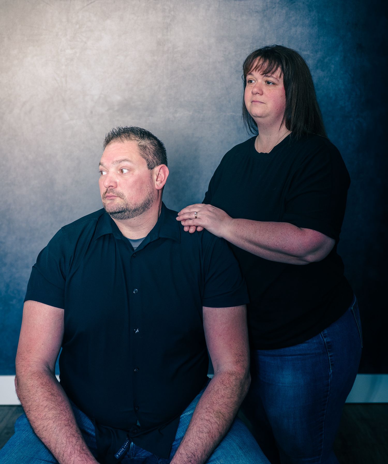 Studio portrait with dramatic lighting showing two people in black clothing against a textured blue-gray background.