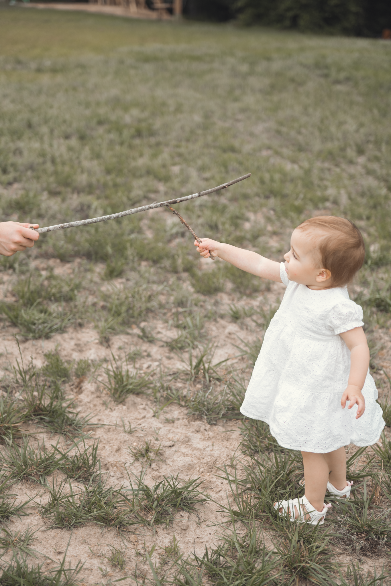 Toddler in white dress holds stick while standing on patchy grass field with another person.