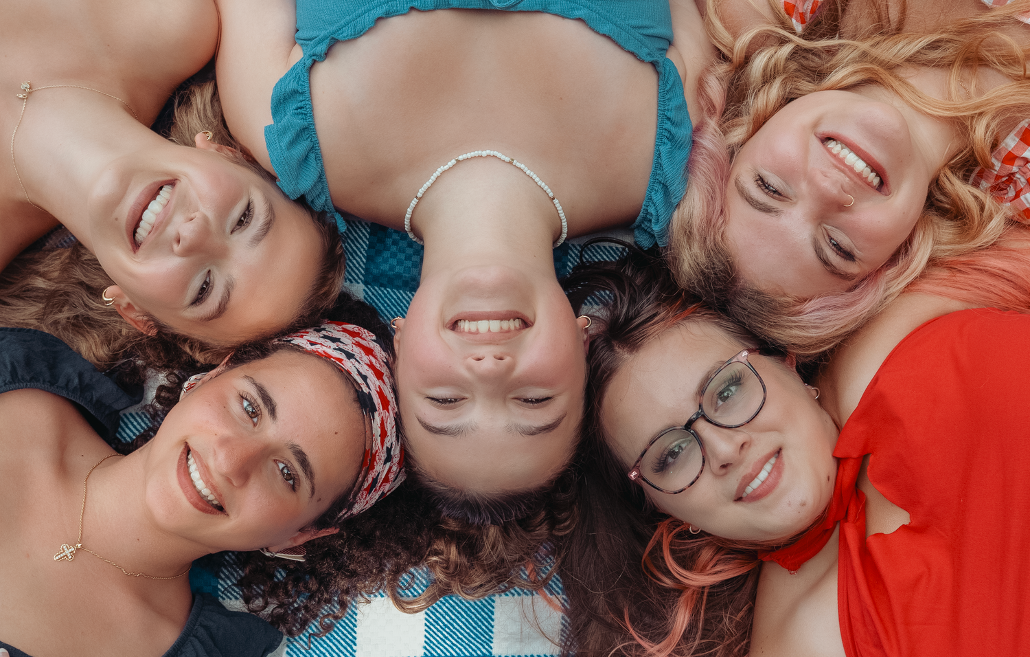 Group of young people lying in circle formation looking down at camera smiling in casual colorful clothing.