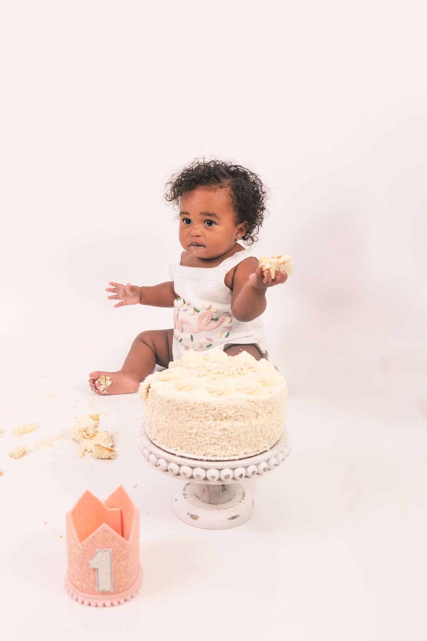 First birthday baby cake smash session with white cake and coral pink crown on light backdrop.