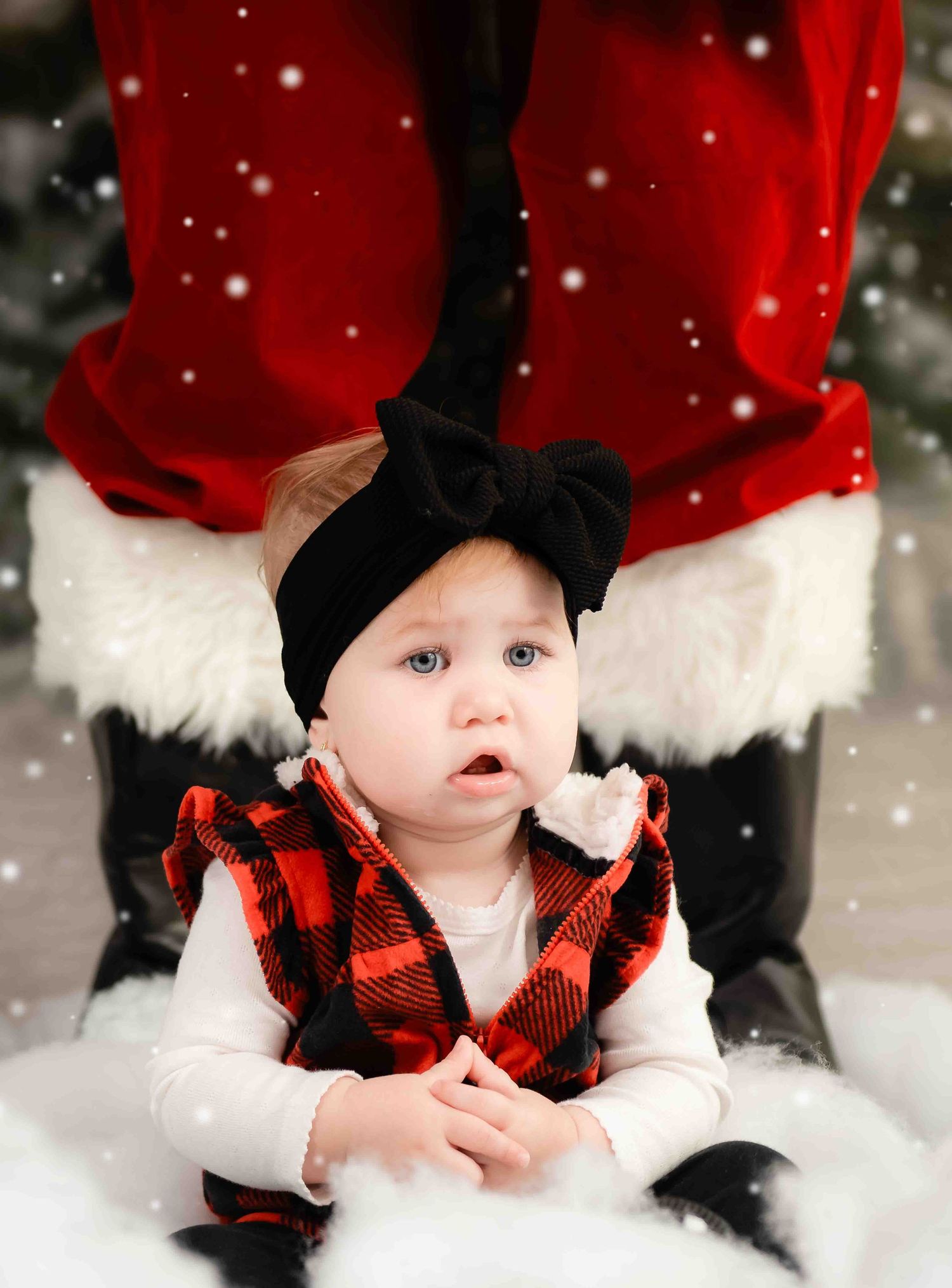 A toddler wearing a buffalo plaid vest and black bow headband sits near festive Santa suit trim in a snowy setting.