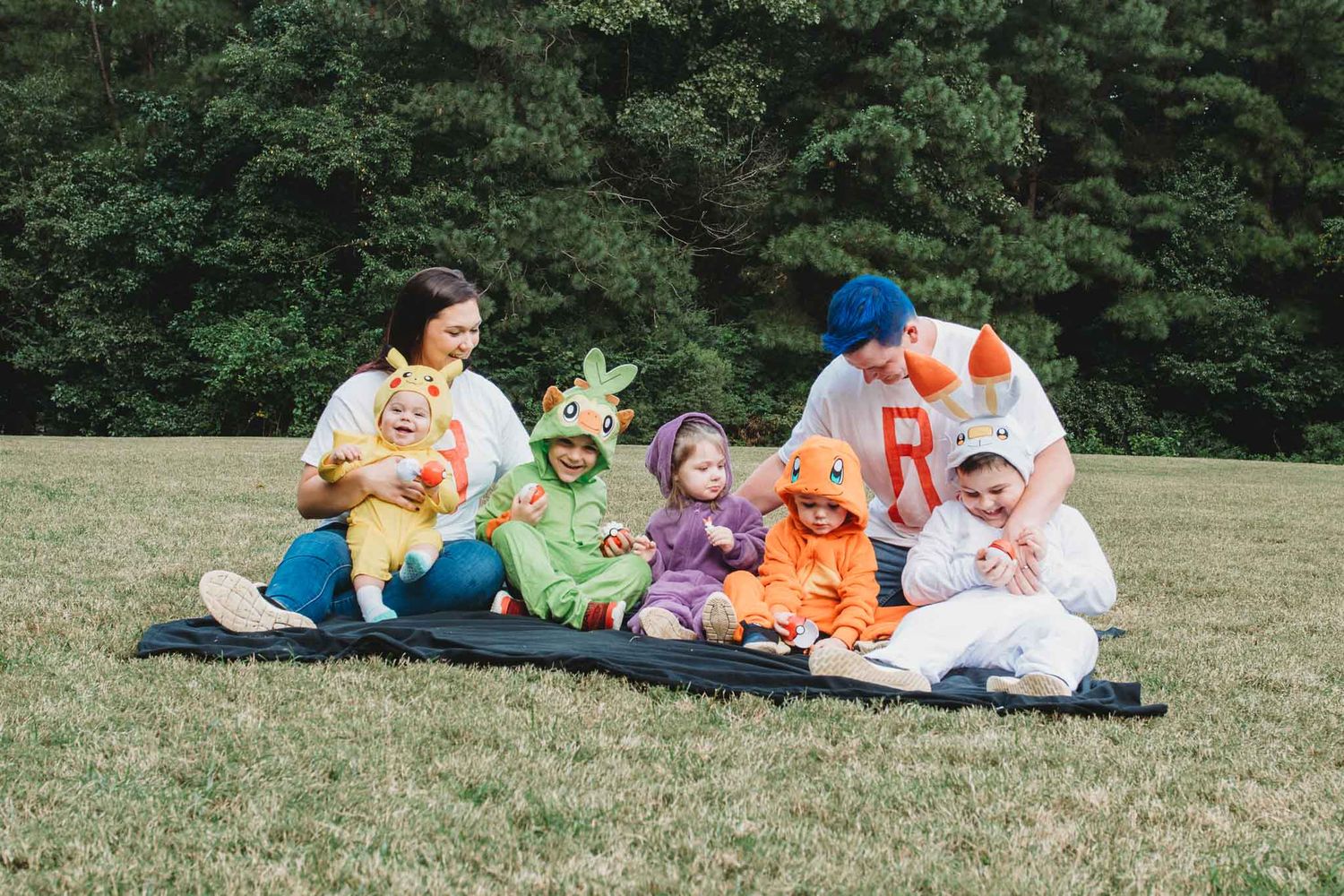 A group sits together on a picnic blanket in a grassy field with trees in the background wearing colorful summer clothing.