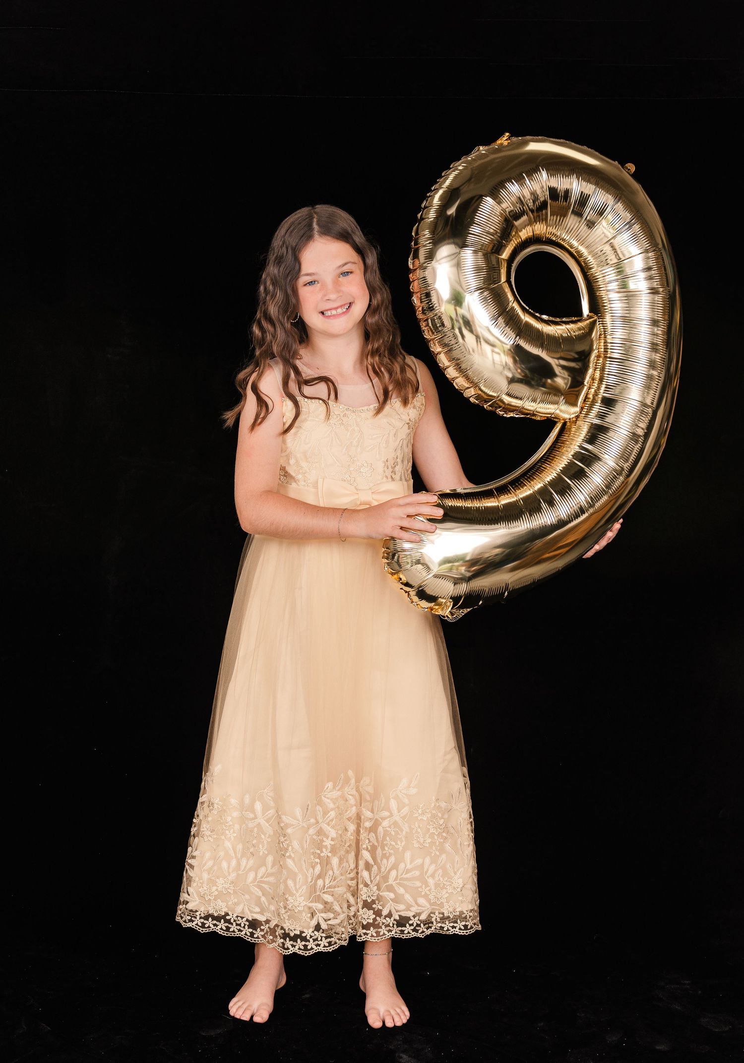 Young girl in cream lace dress holds large gold number 9 balloon against black background in birthday photo.