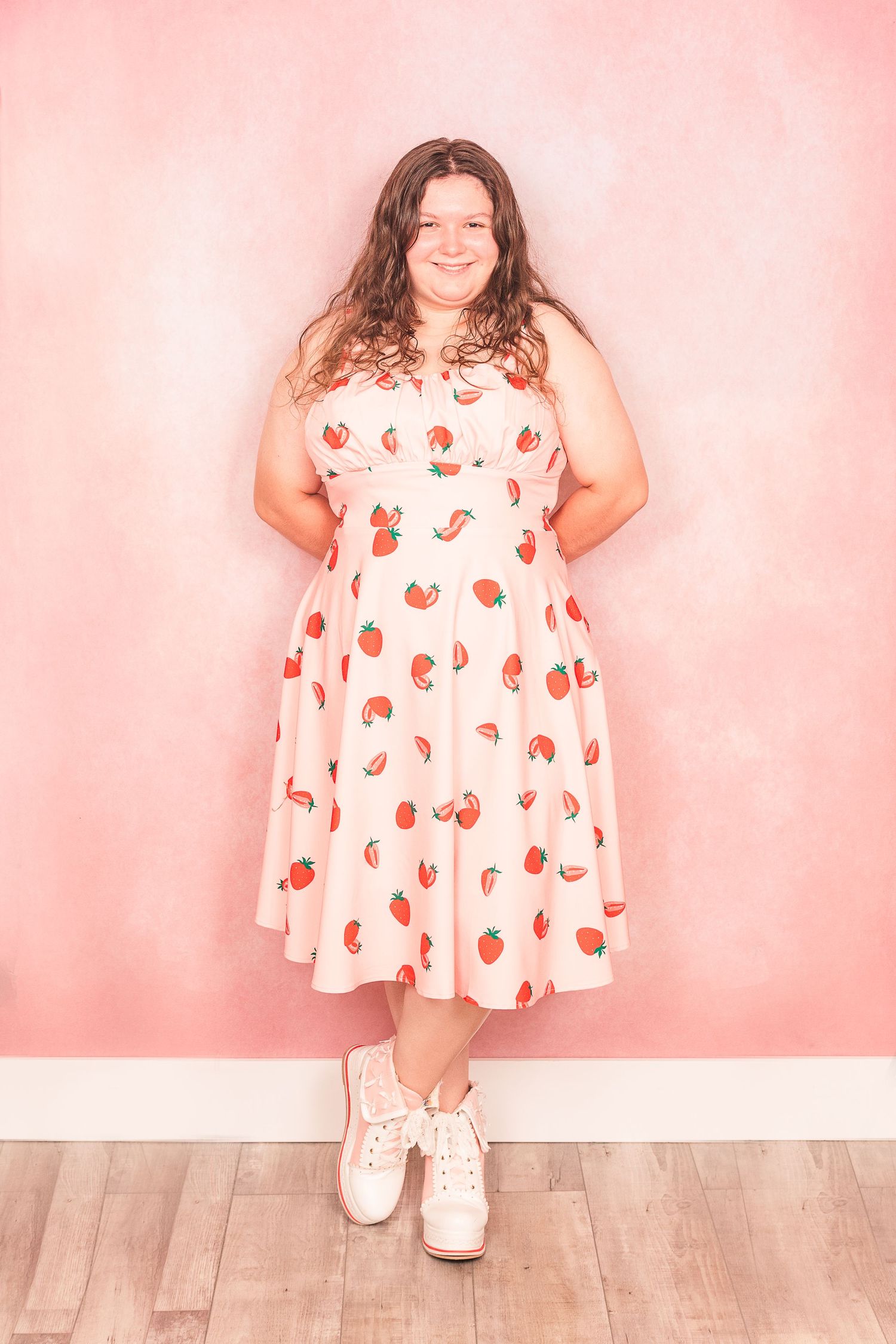 A person in a white dress with cherry print poses against a pink wall wearing white sneakers.