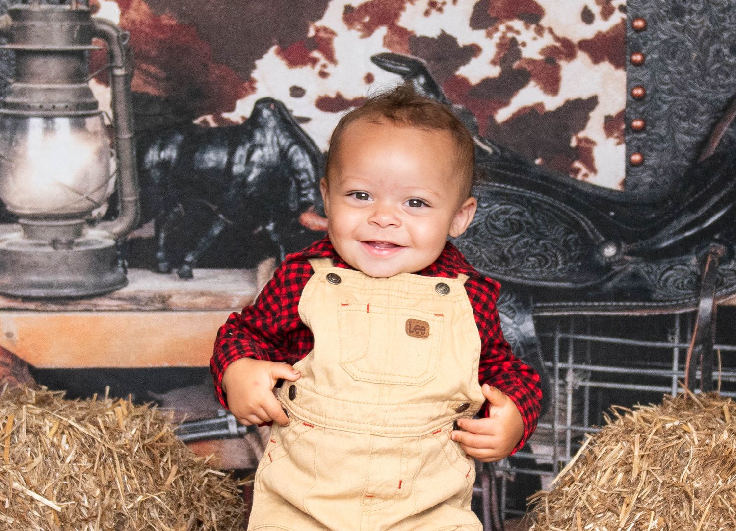A child in plaid and overalls poses near rustic lanterns and hay bales against a patterned backdrop.